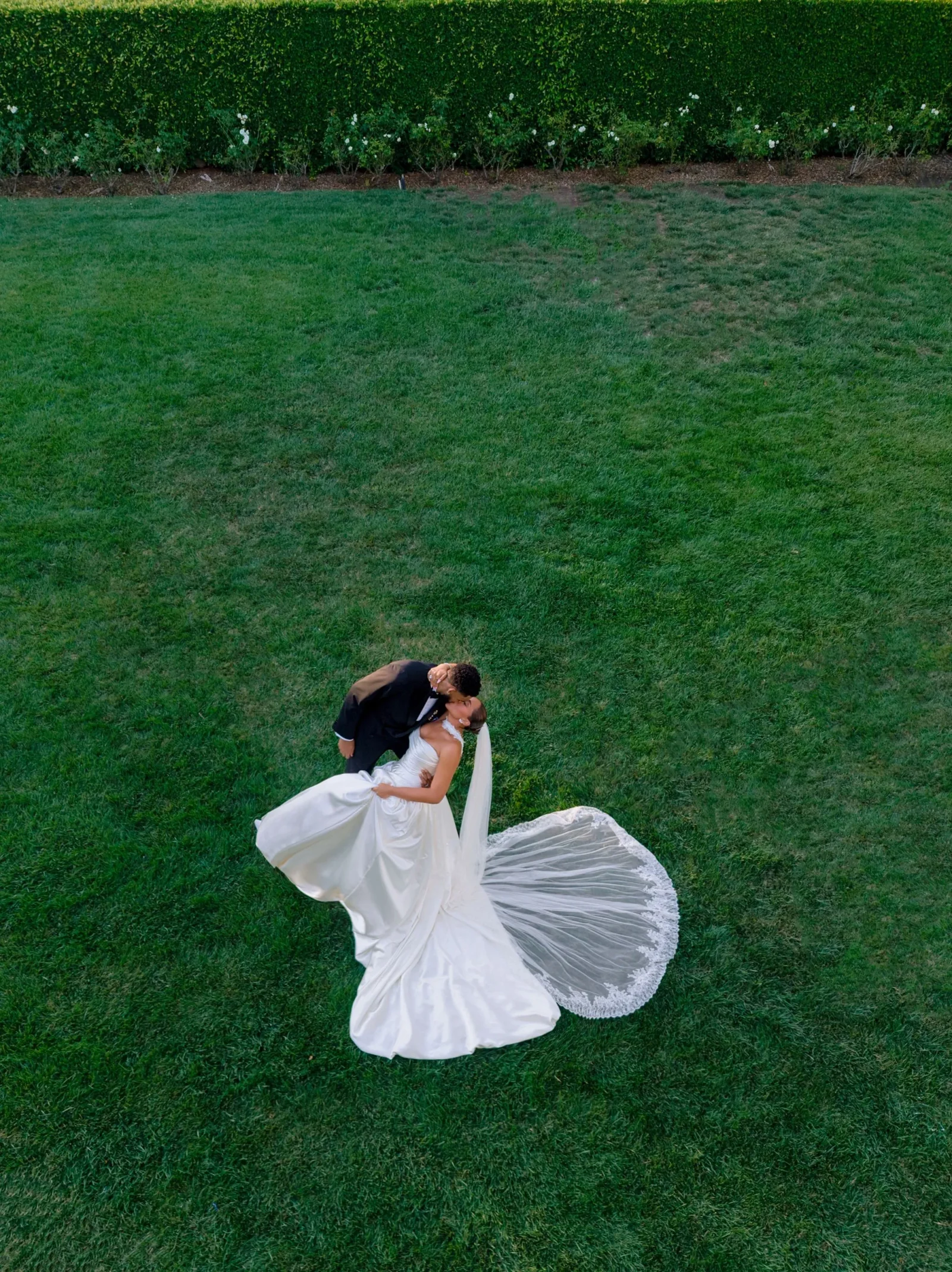 Aerial view of a bride in a white wedding dress and a groom in a dark suit kissing on a green lawn, dress train flowing around them.