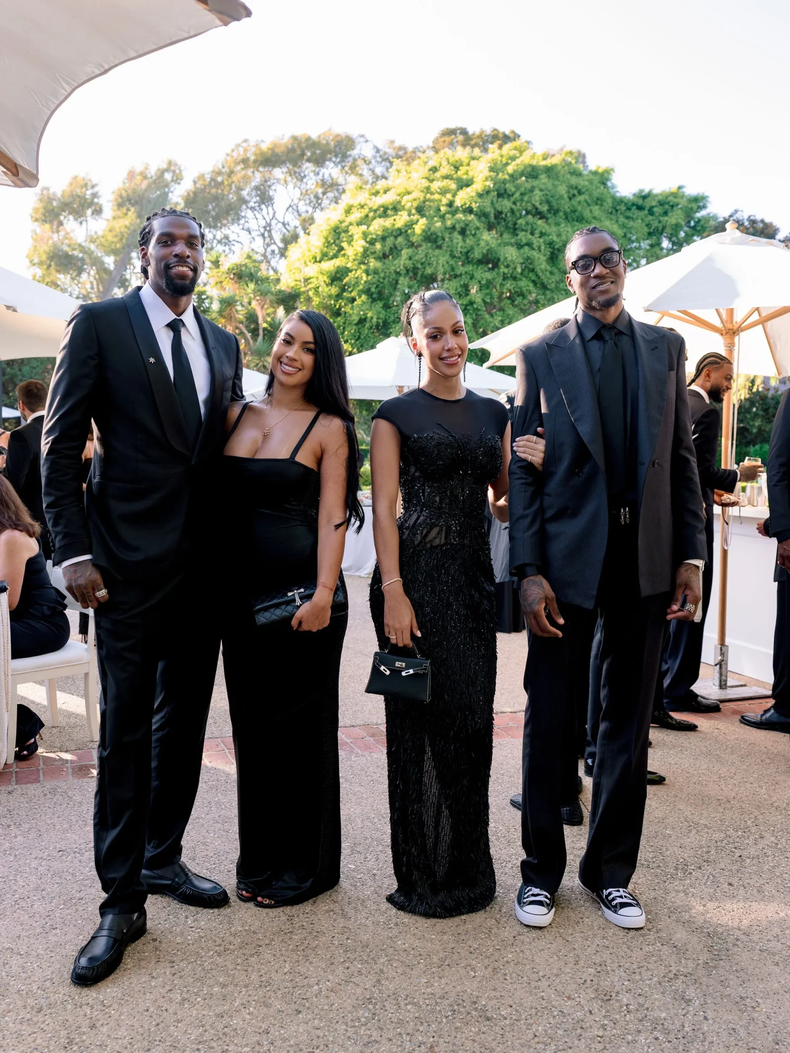 Group of four adults in black formalwear posing outdoors at a garden event, with white tents and green trees in the background.