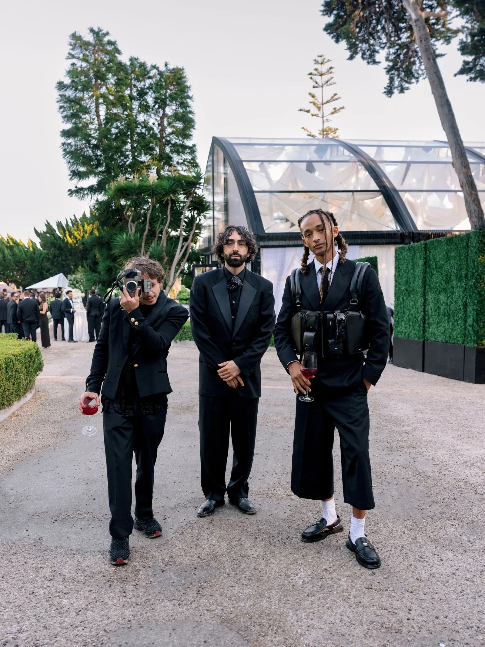 Three men in black suits posing outdoors at a formal event, one holding a camera and wine glass near a glass-domed structure.