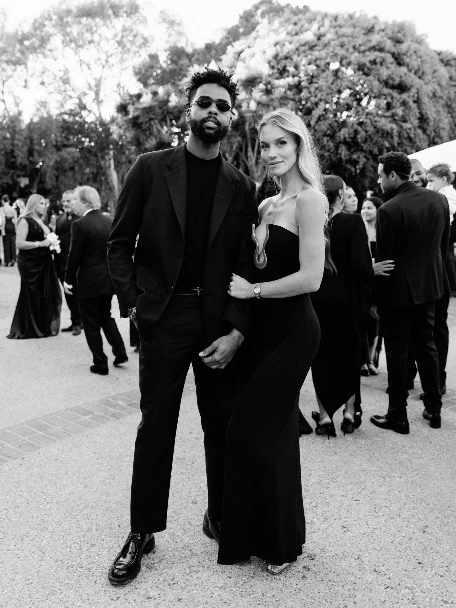 Couple in formal black attire posing together at an outdoor gala with guests in the background (black-and-white).