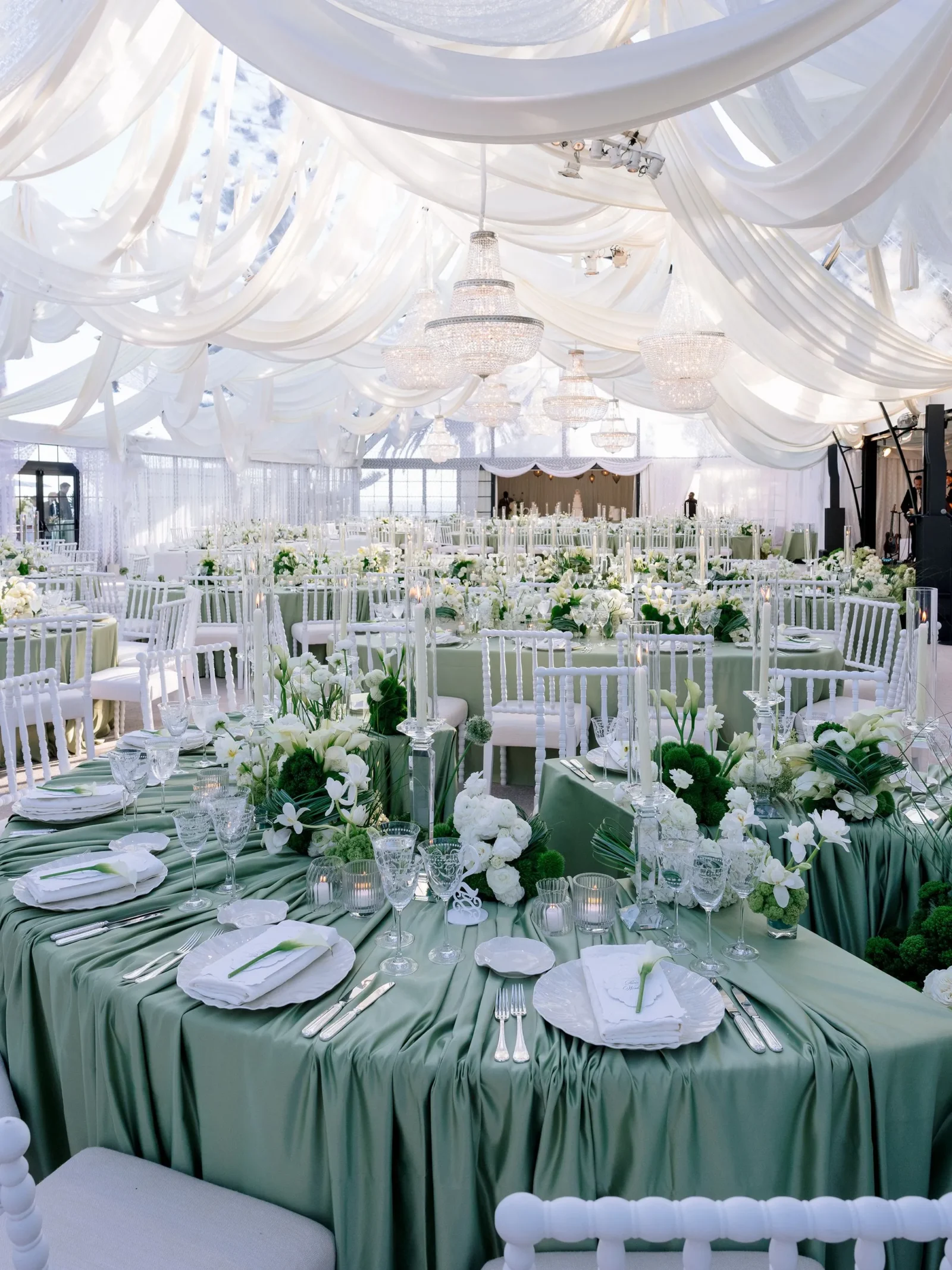 Wedding reception setup with long green-draped tables, white chairs, and floral centerpieces under flowing white fabric and crystal chandeliers.