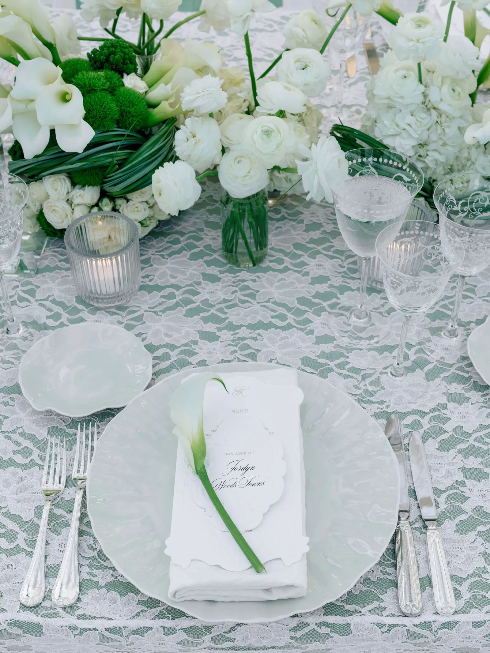 Elegant white-themed wedding table with a floral centerpiece, crystal glasses, and a menu card on a lace tablecloth.