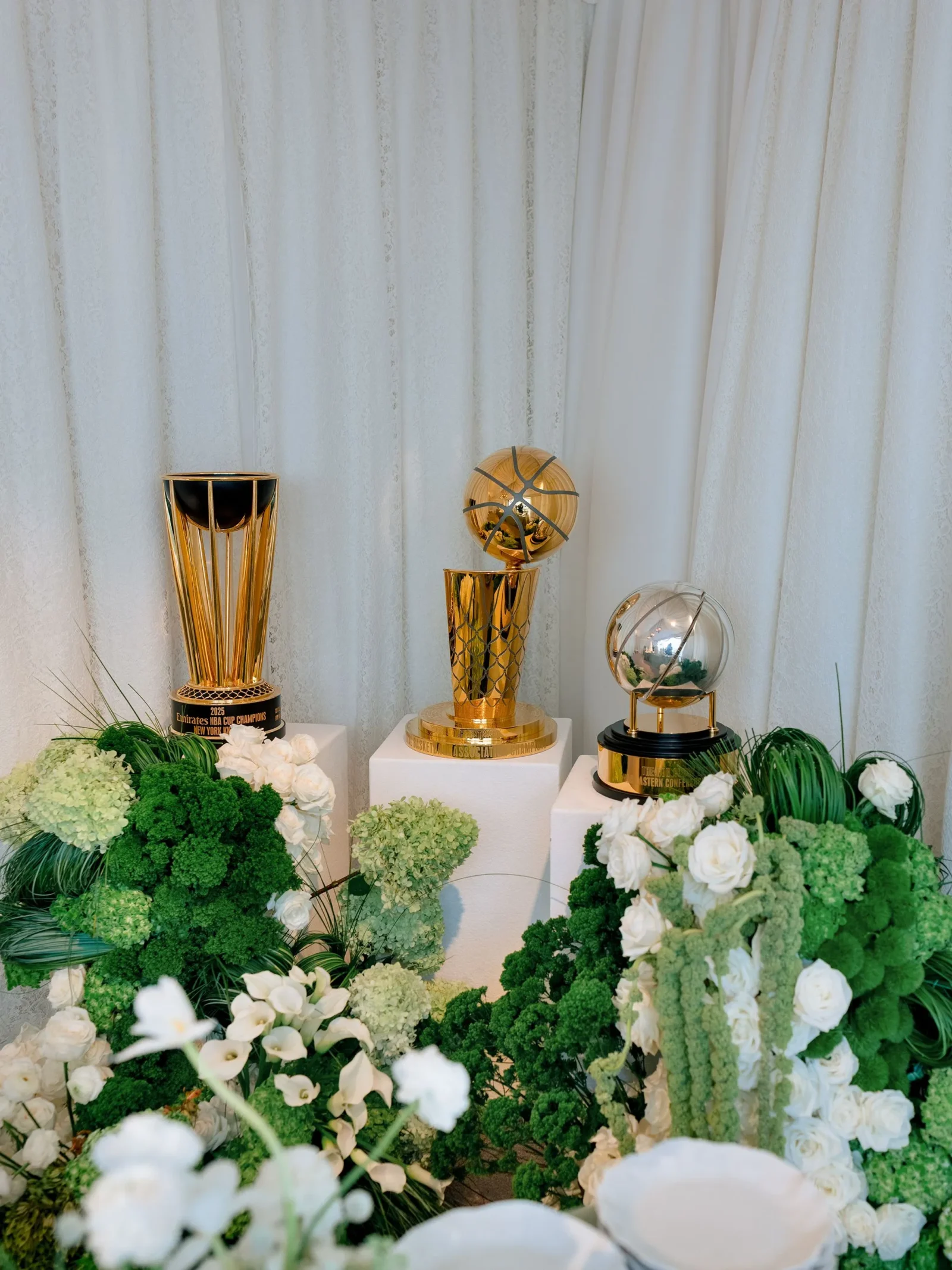 Gold trophy on a pedestal with green-and-white floral arrangements in front, against a white draped backdrop.