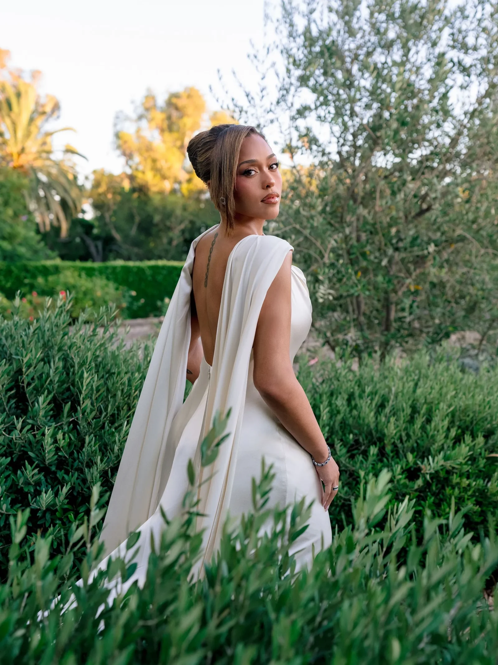 Woman in a backless white dress standing in a lush garden, looking over her shoulder toward the camera.