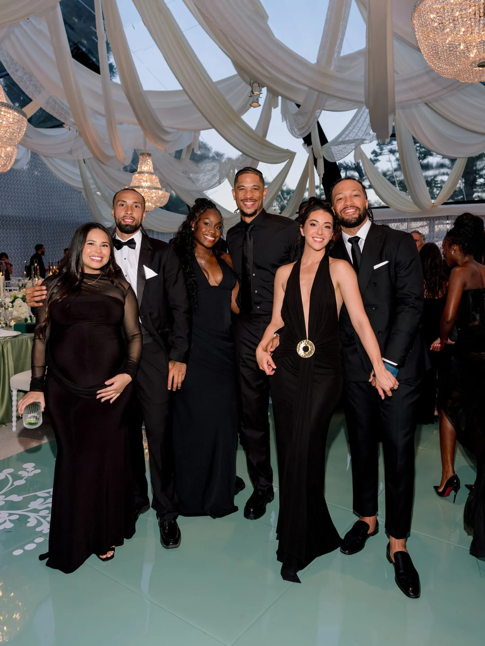 Group of six friends in formal black attire posing together at an elegant indoor event with chandeliers and white draped fabric overhead.
