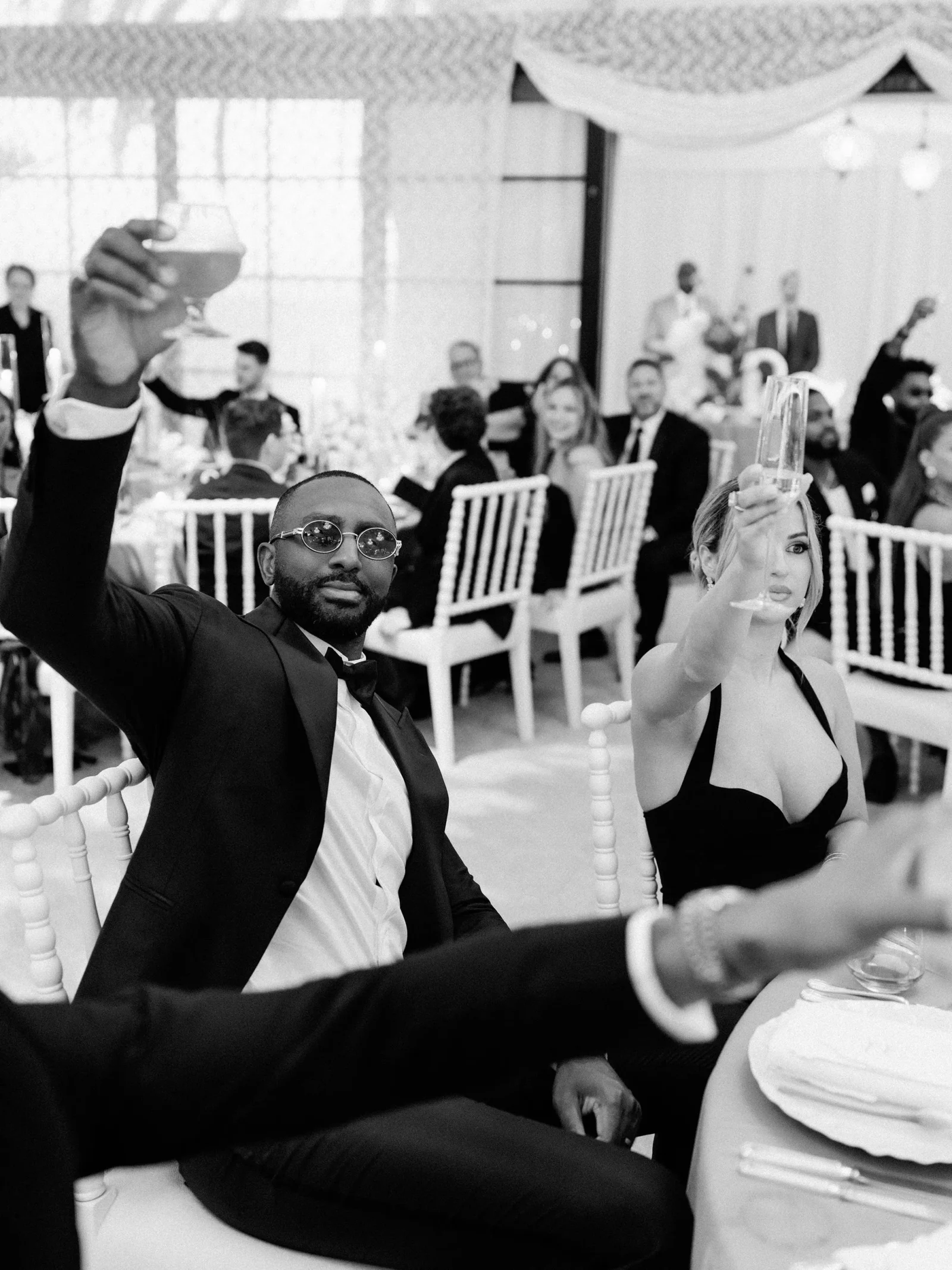 Man in a tuxedo raises a glass at a wedding reception while a woman beside him toasts; guests mingle in the background in black-tie attire.