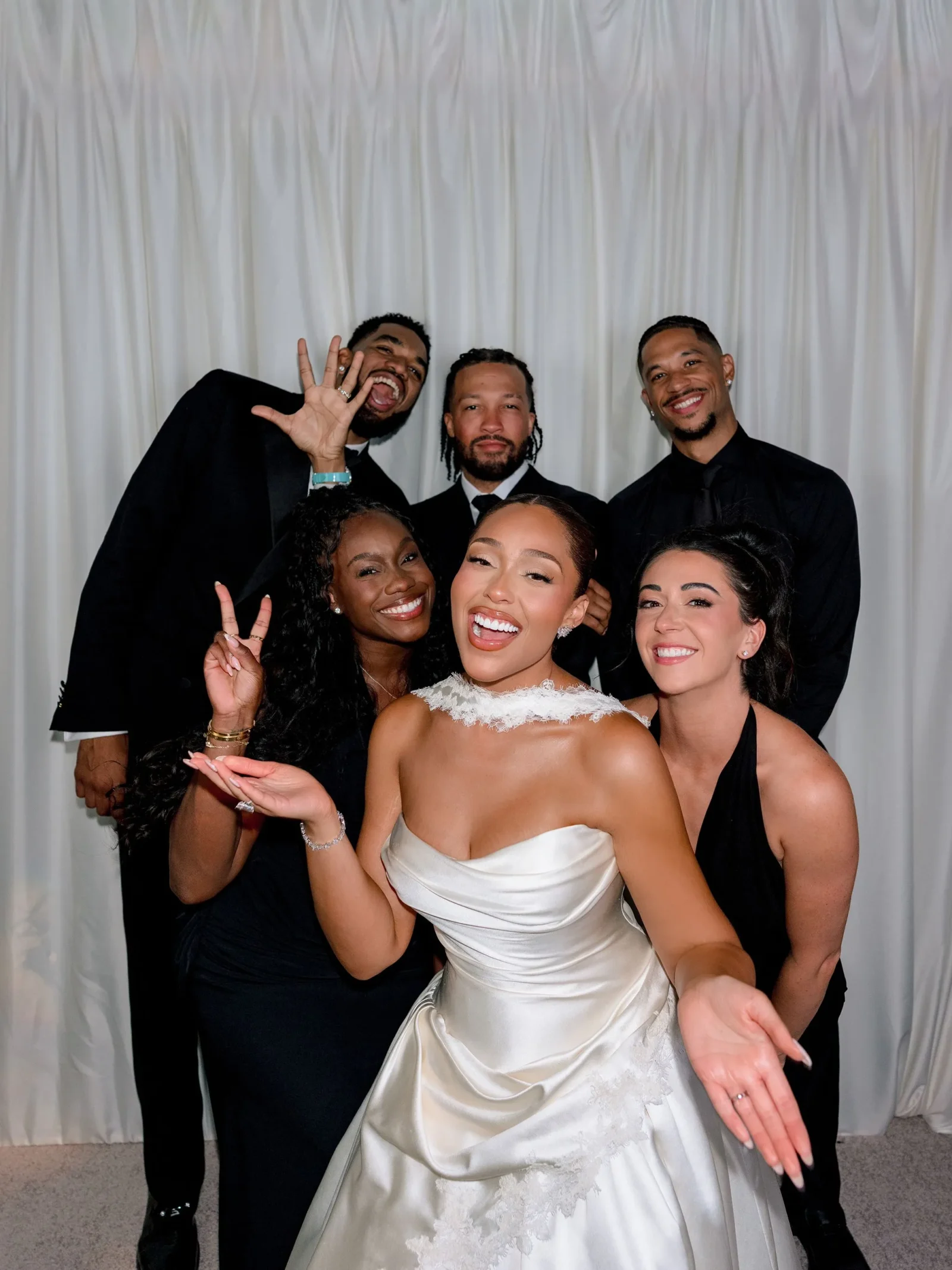 Bride in a white gown front and center, flanked by two smiling women and three men in black suits posing in front of a white curtain backdrop.
