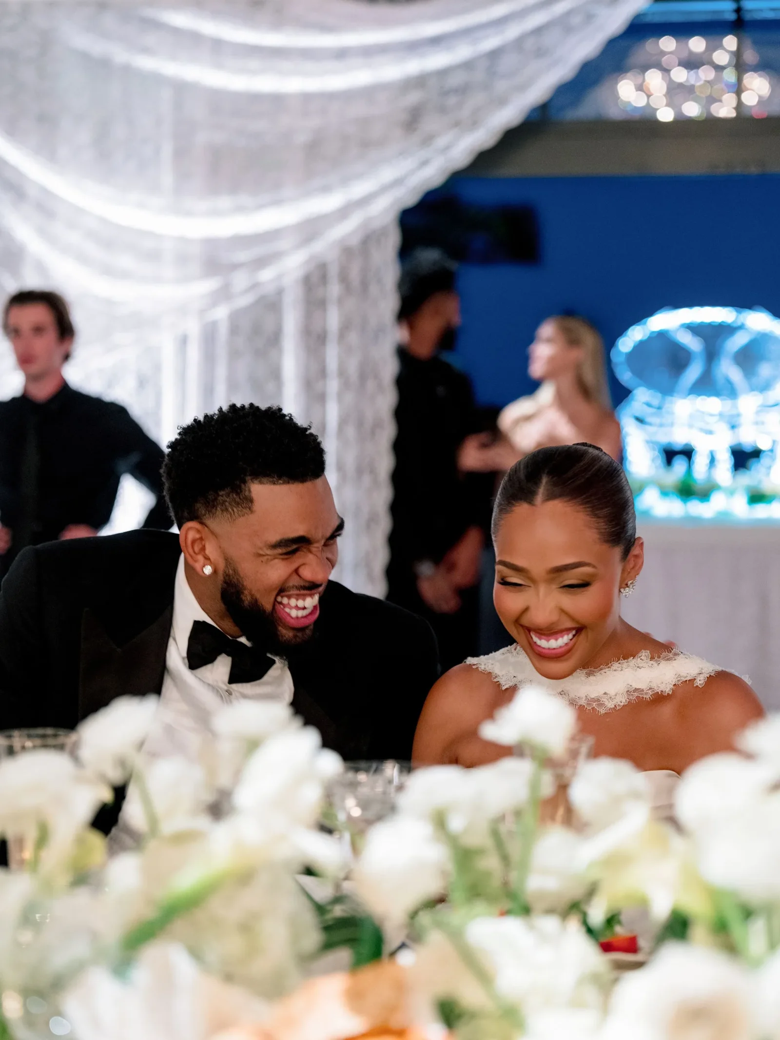A laughing couple in formal attire seated at a wedding reception, foreground flowers blur the scene