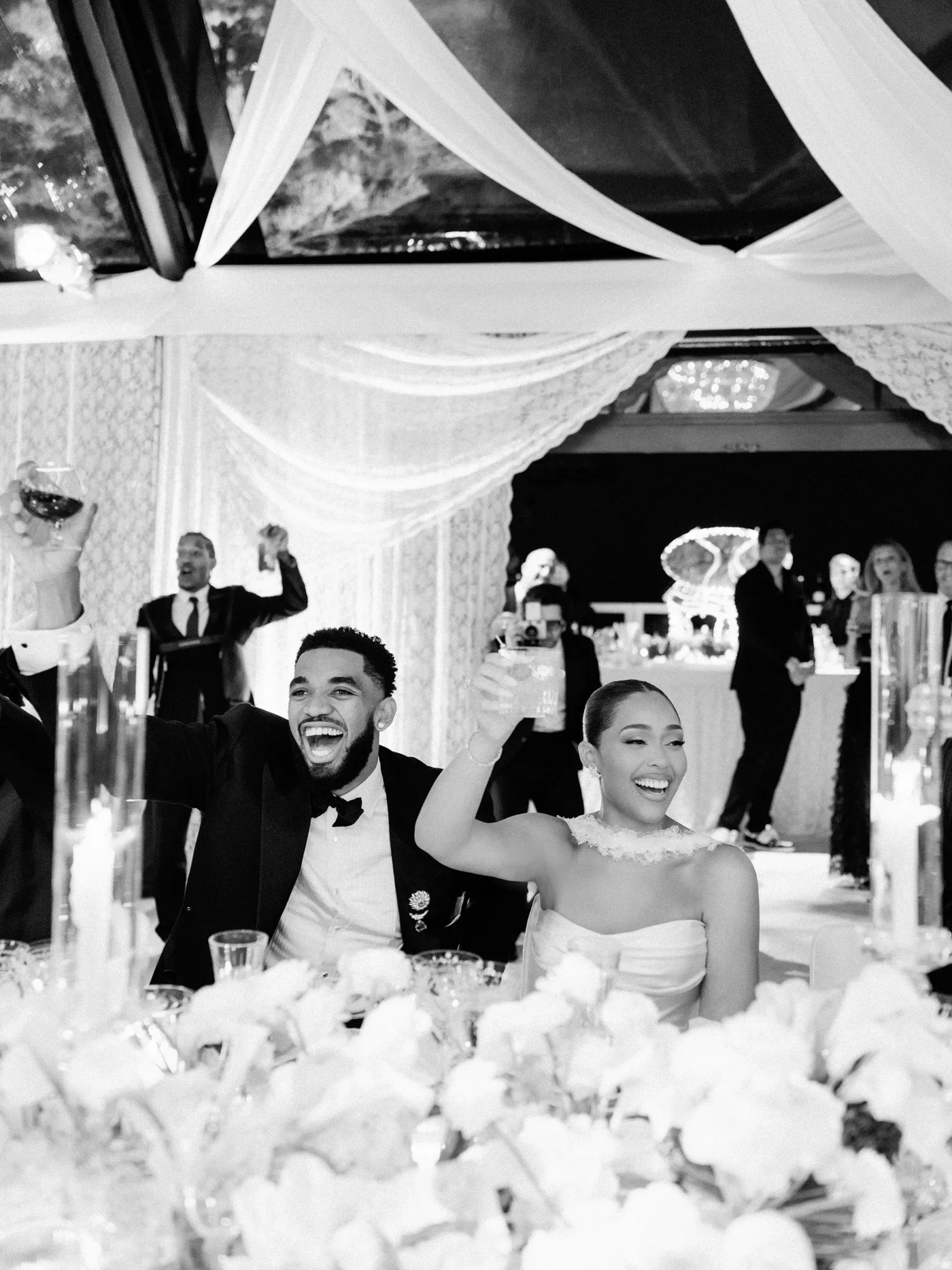 Bride and groom raise glasses in a toast at a decorated wedding reception, with guests and flowers in the foreground.