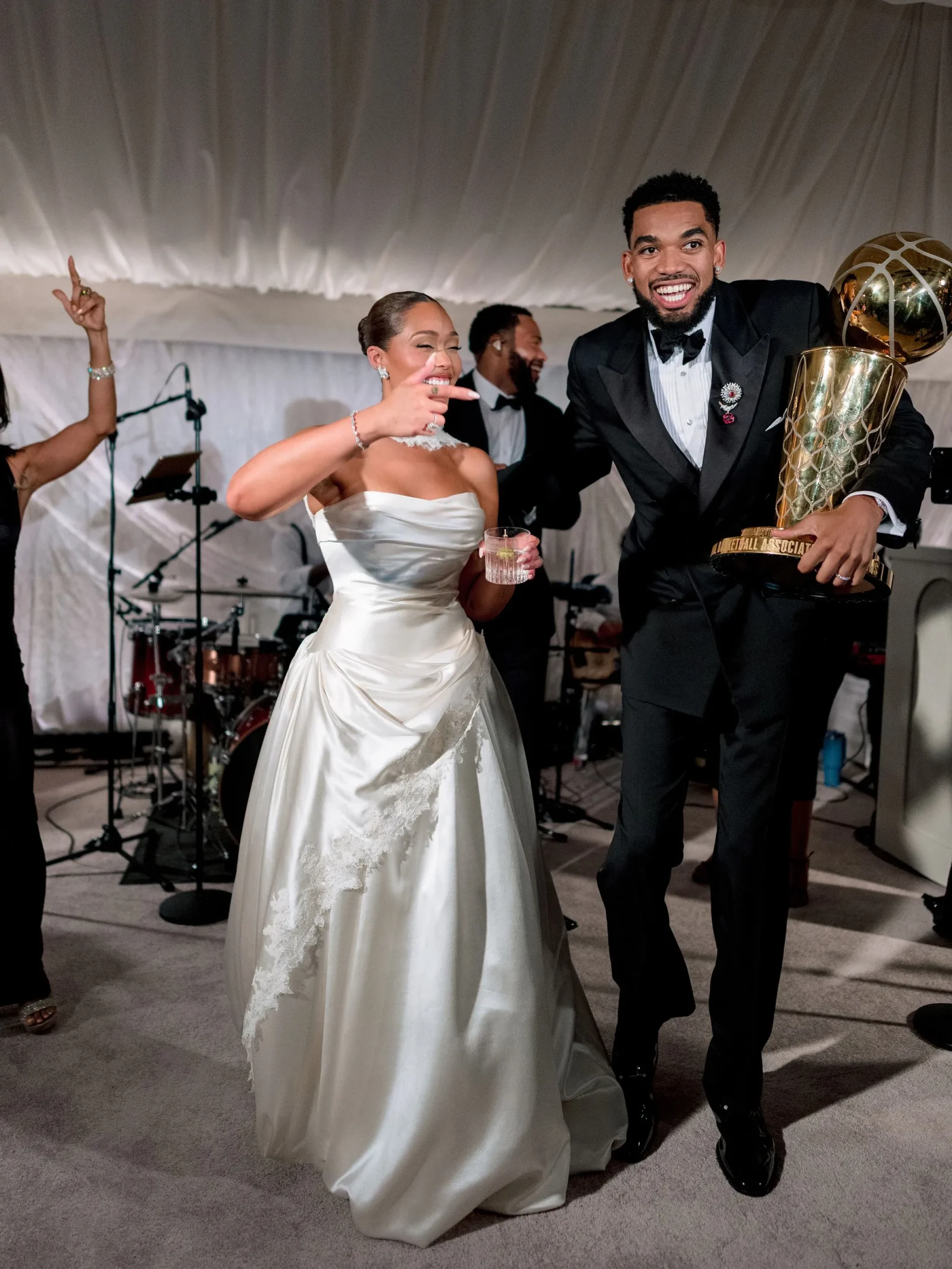 Bride in a white wedding dress and a smiling man in a tuxedo pose with a large gold trophy at a live band reception stage in the background.