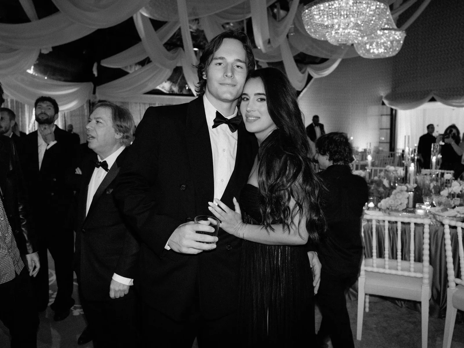Couple in formal attire posing at a black-tie gala, man in a tuxedo and bow tie, woman in a black dress holding a drink, chandeliers above.