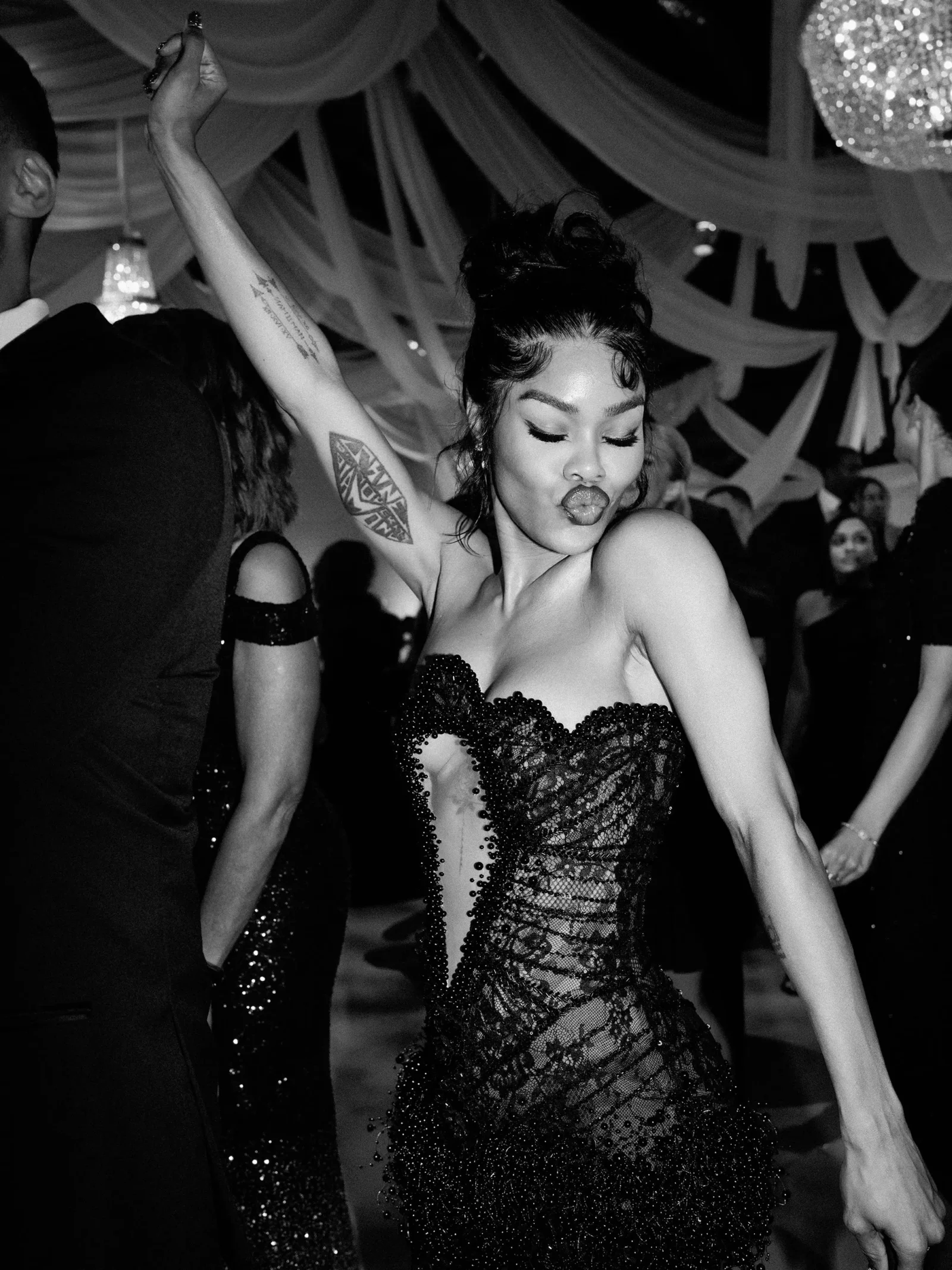 Woman dancing at a party with one arm raised, wearing a sparkly lace dress and showing a tattoo on her forearm while puckering her lips.