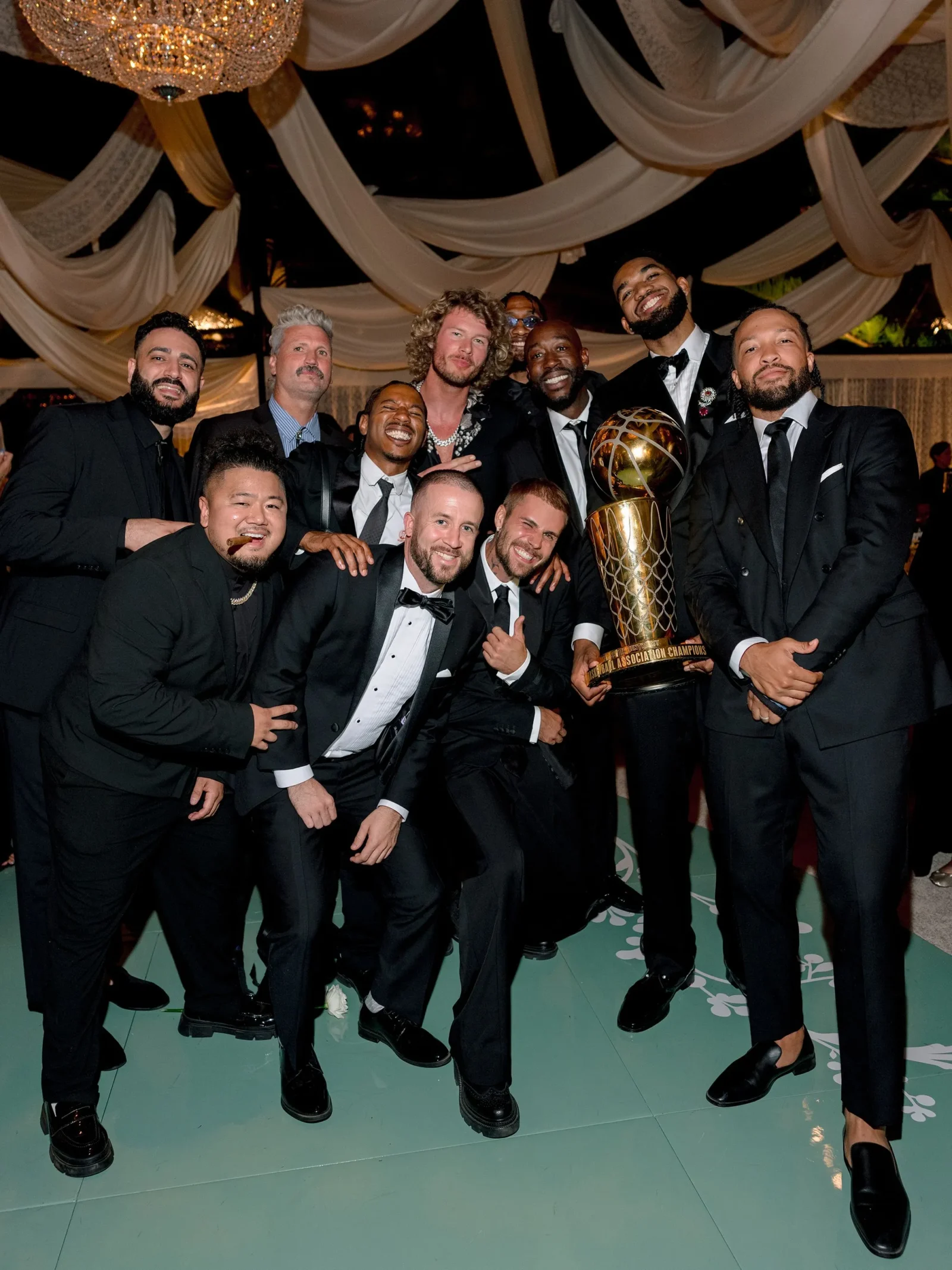 Group of men in black tuxedos posing with a tall gold trophy at a formal event under draped ceilings and chandeliers.
