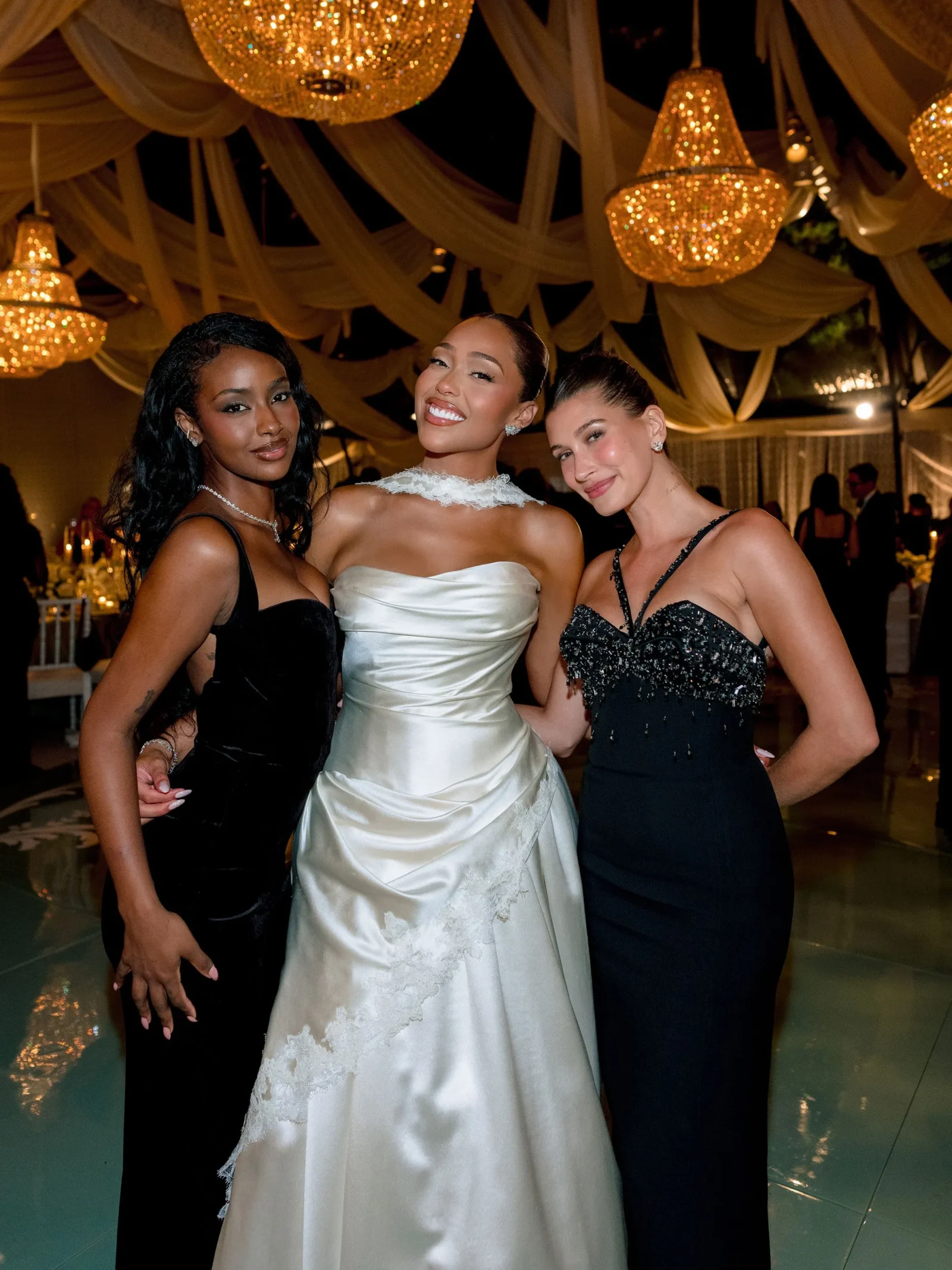Three women in formal gowns posing at a glamorous event with chandeliers in the background; center wears a white wedding-style dress.