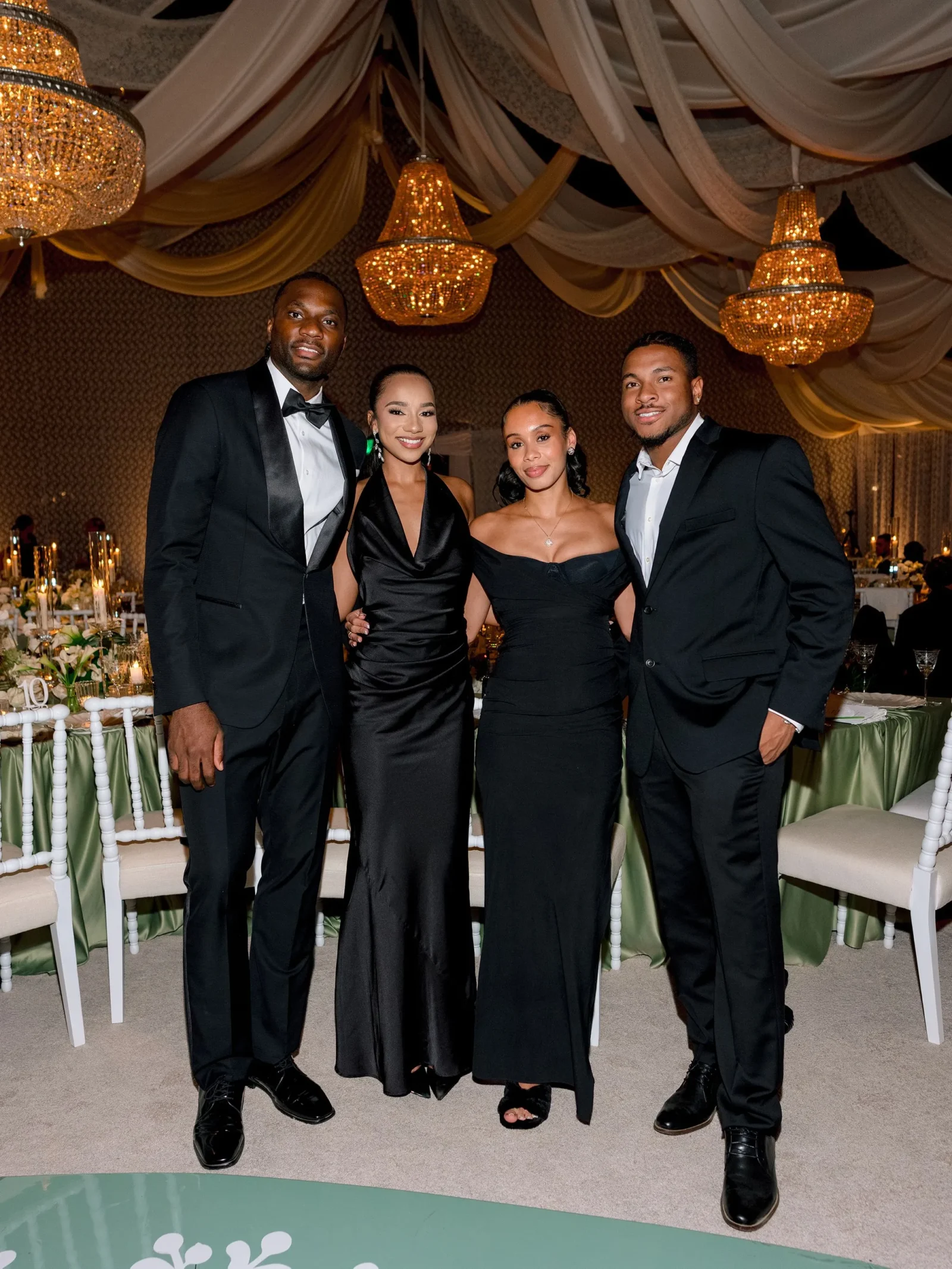Four formally dressed friends posing together at a banquet hall with chandeliers and draped ceilings; two men in black suits and two women in black gowns smile for the camera.