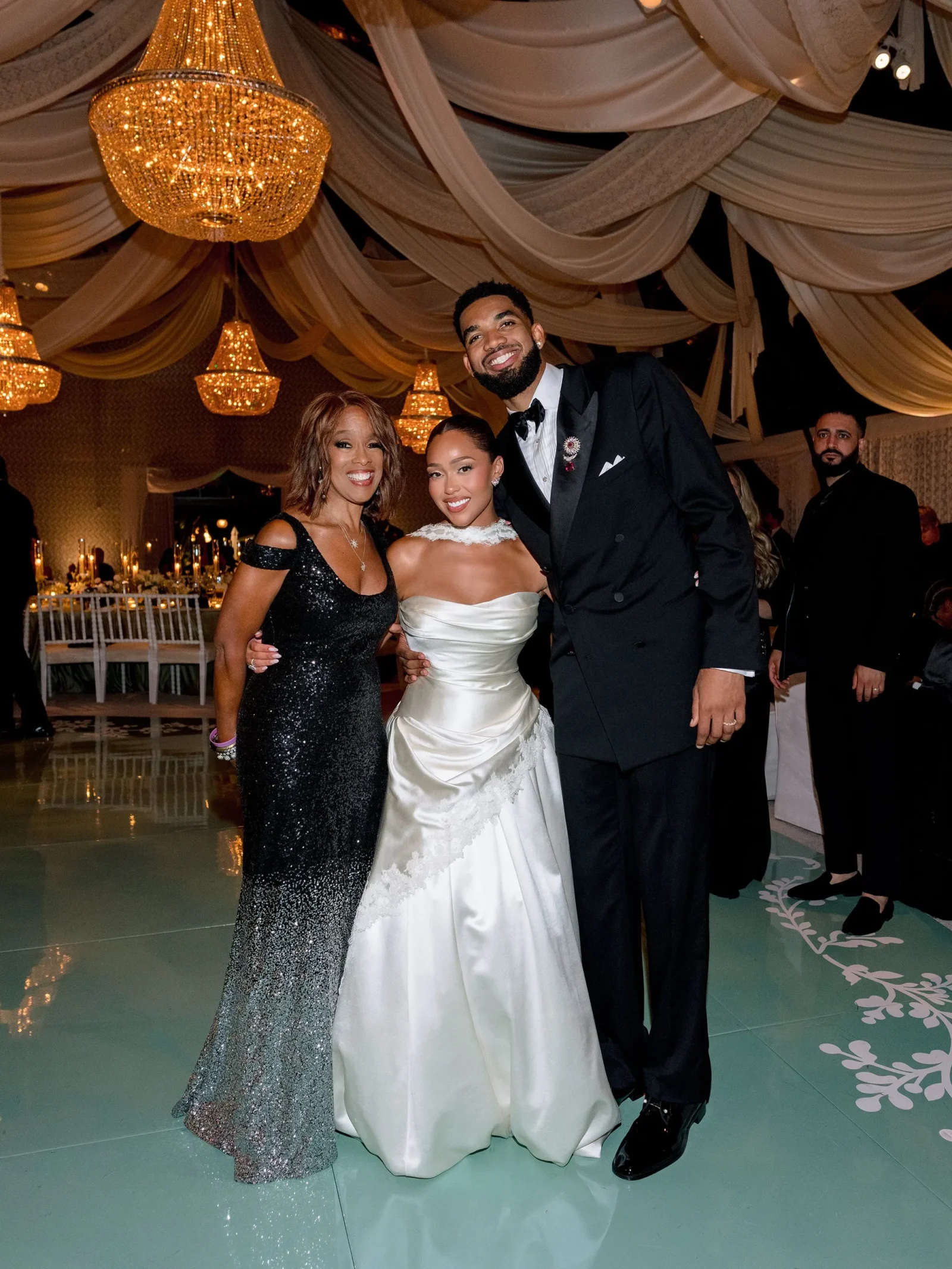 Bride in a white gown posing with a man in a black tuxedo and a woman in a sequined black dress at a formal wedding reception under large chandeliers.