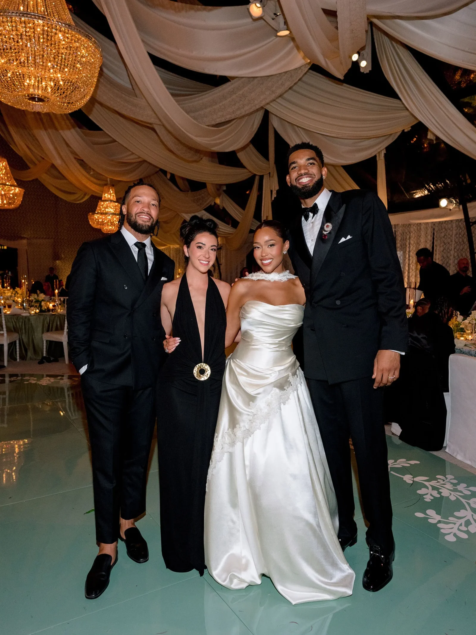 Group of four in formal wear posing and smiling at a wedding reception with chandeliers and draped ceiling in the background.