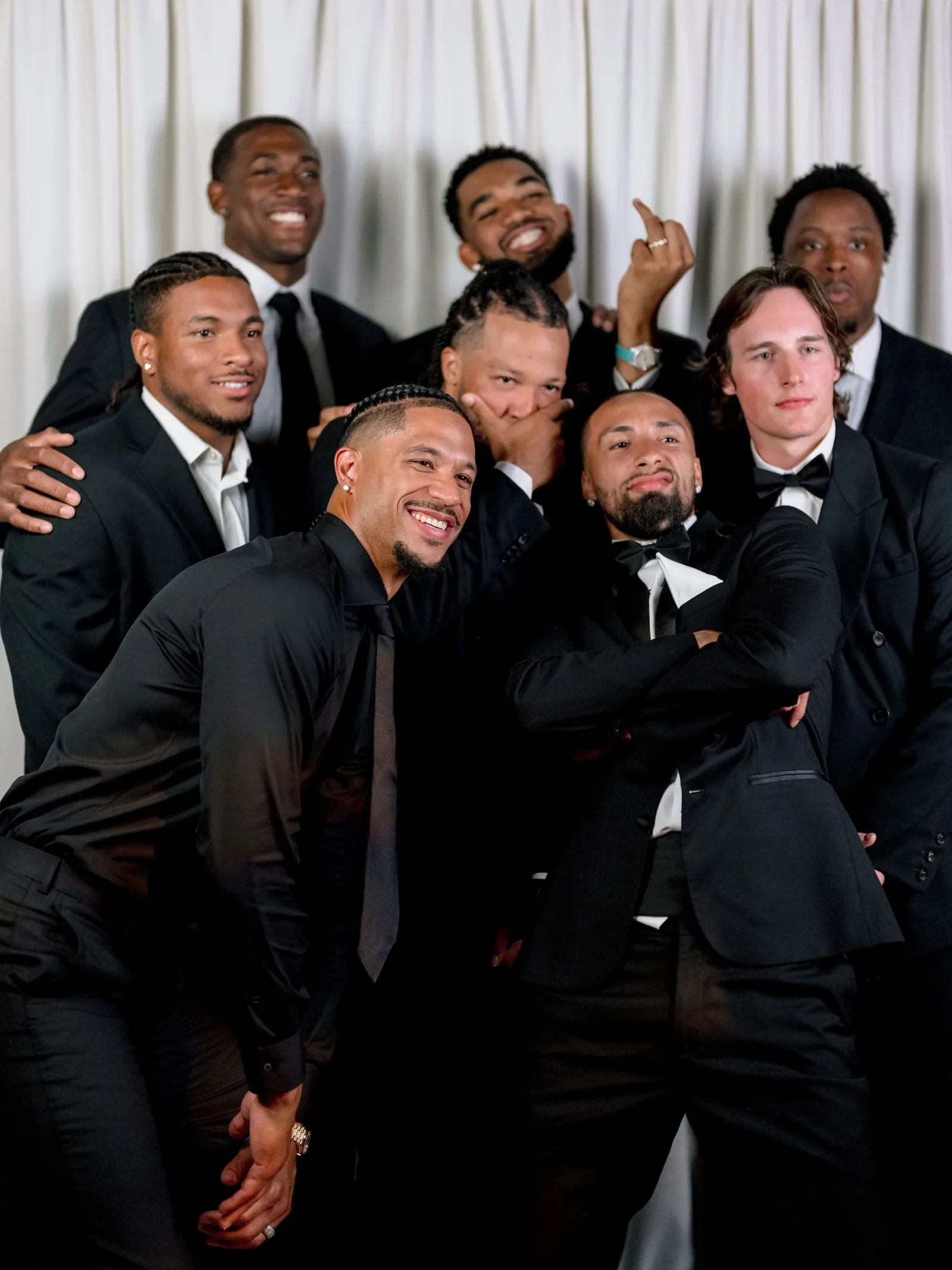 Group of men in black tuxedos posing together for a photo, some smiling and making playful gestures in front of a light curtain backdrop.