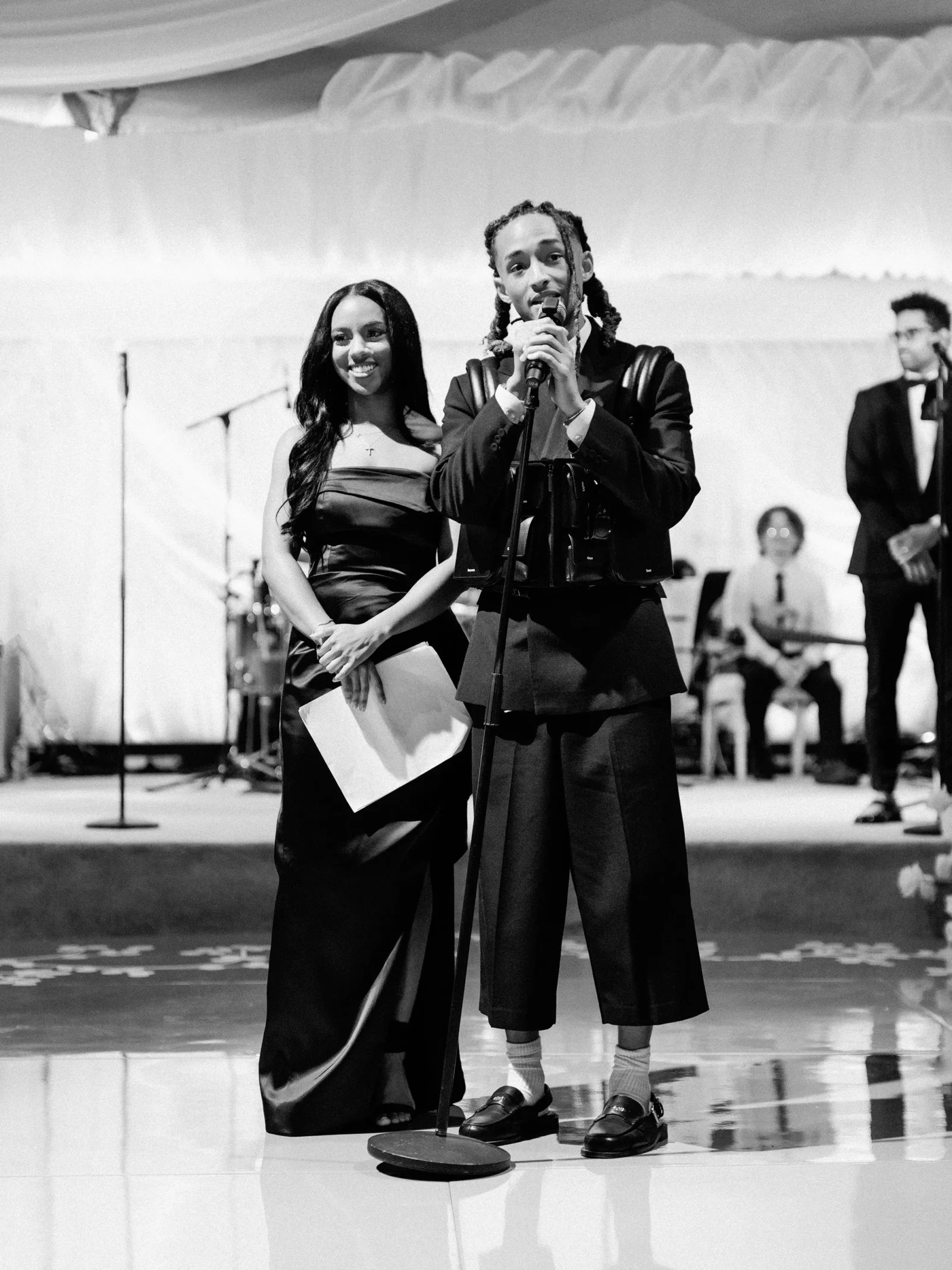 Two performers on a stage: a man sings into a microphone while a smiling woman in a formal dress stands beside him with a paper.