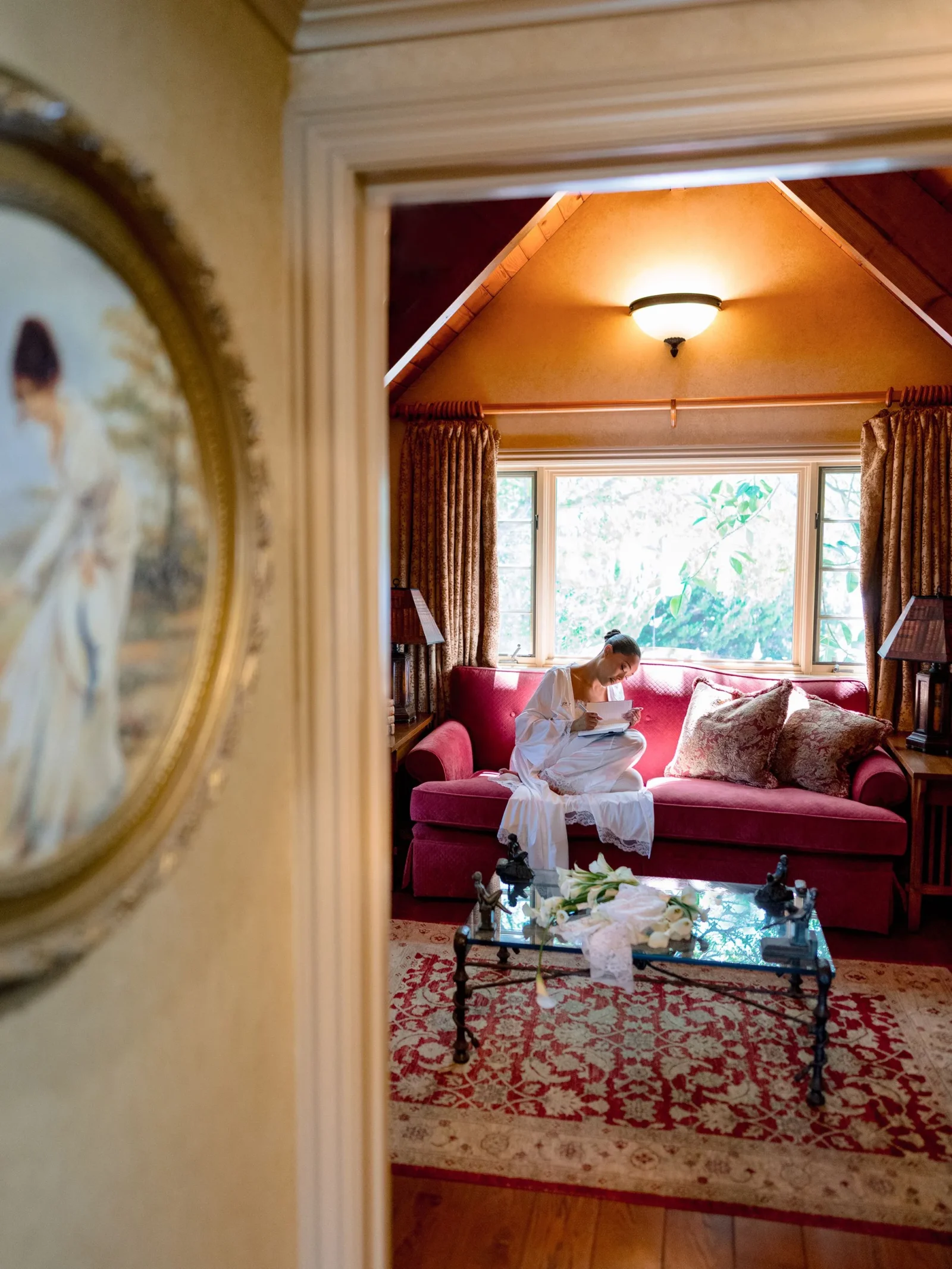 Woman in a white dress sits on a red sofa, reading a book in a sunlit, cozy living room with patterned rug and large window.