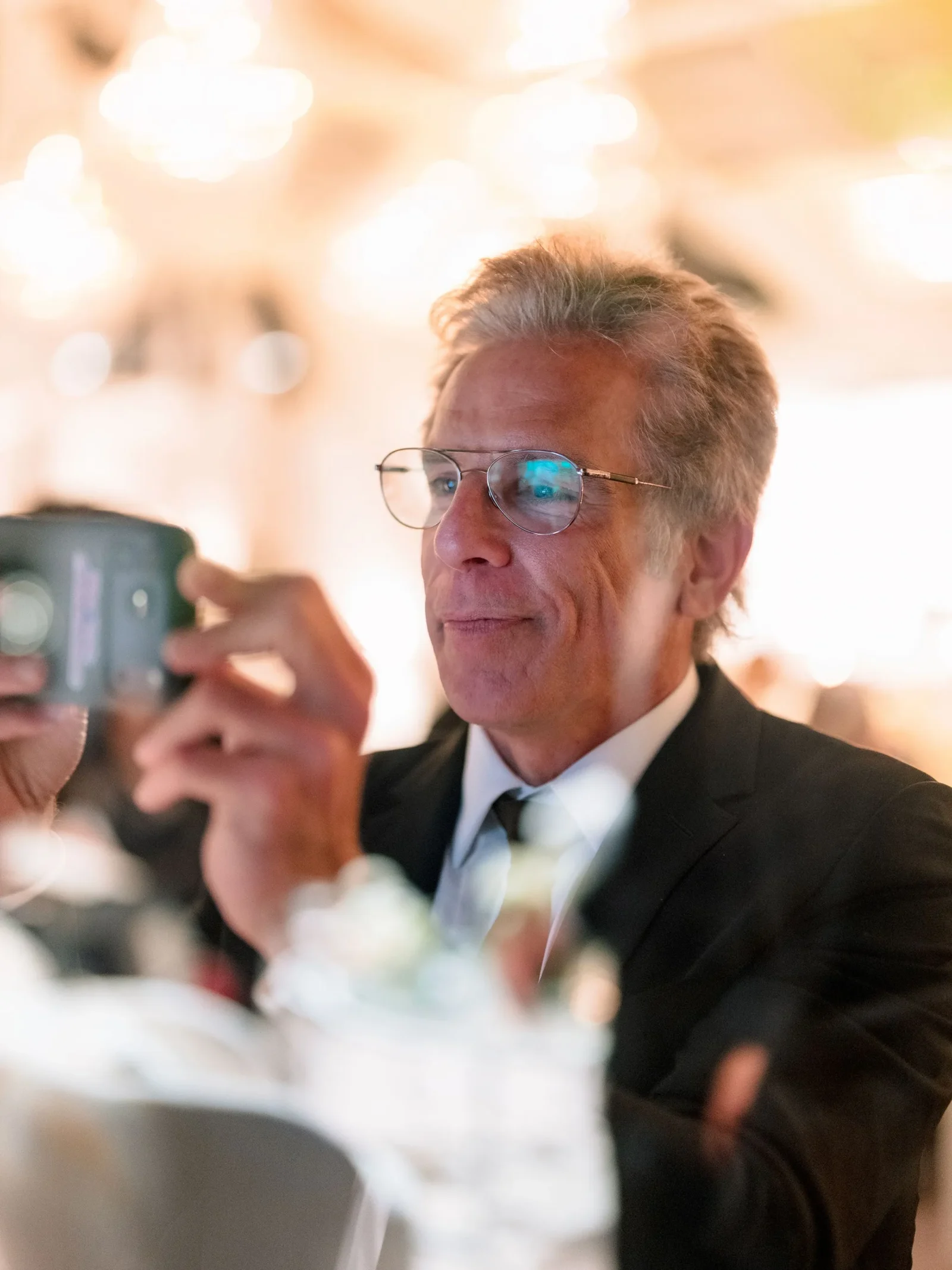 Middle-aged man in a black suit and tie with round glasses, taking a photo with a smartphone at a formal event, warm bokeh lights in the background.