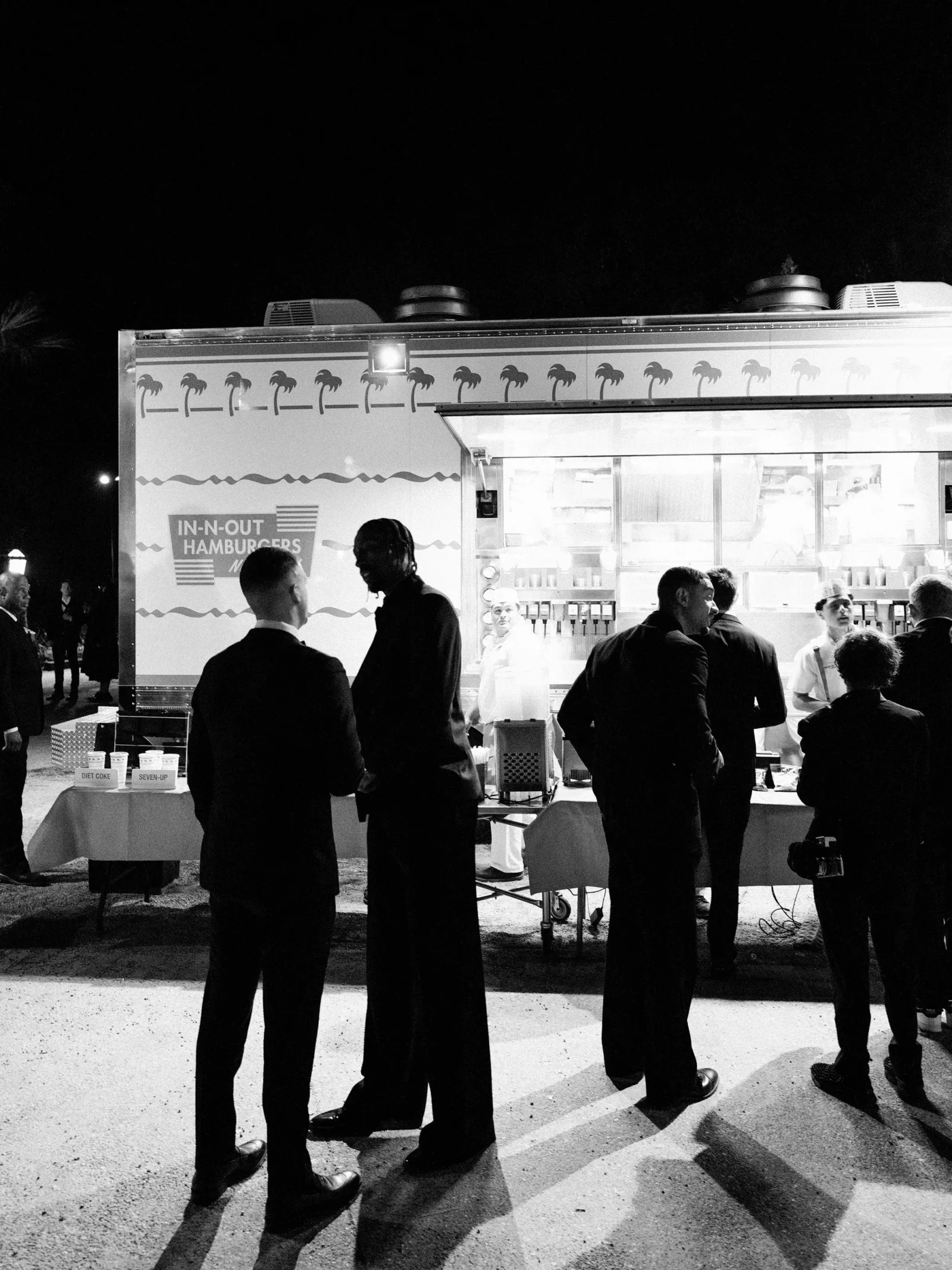Night scene of a palm-decorated food truck with a serving window; silhouettes of people in suits stand nearby.