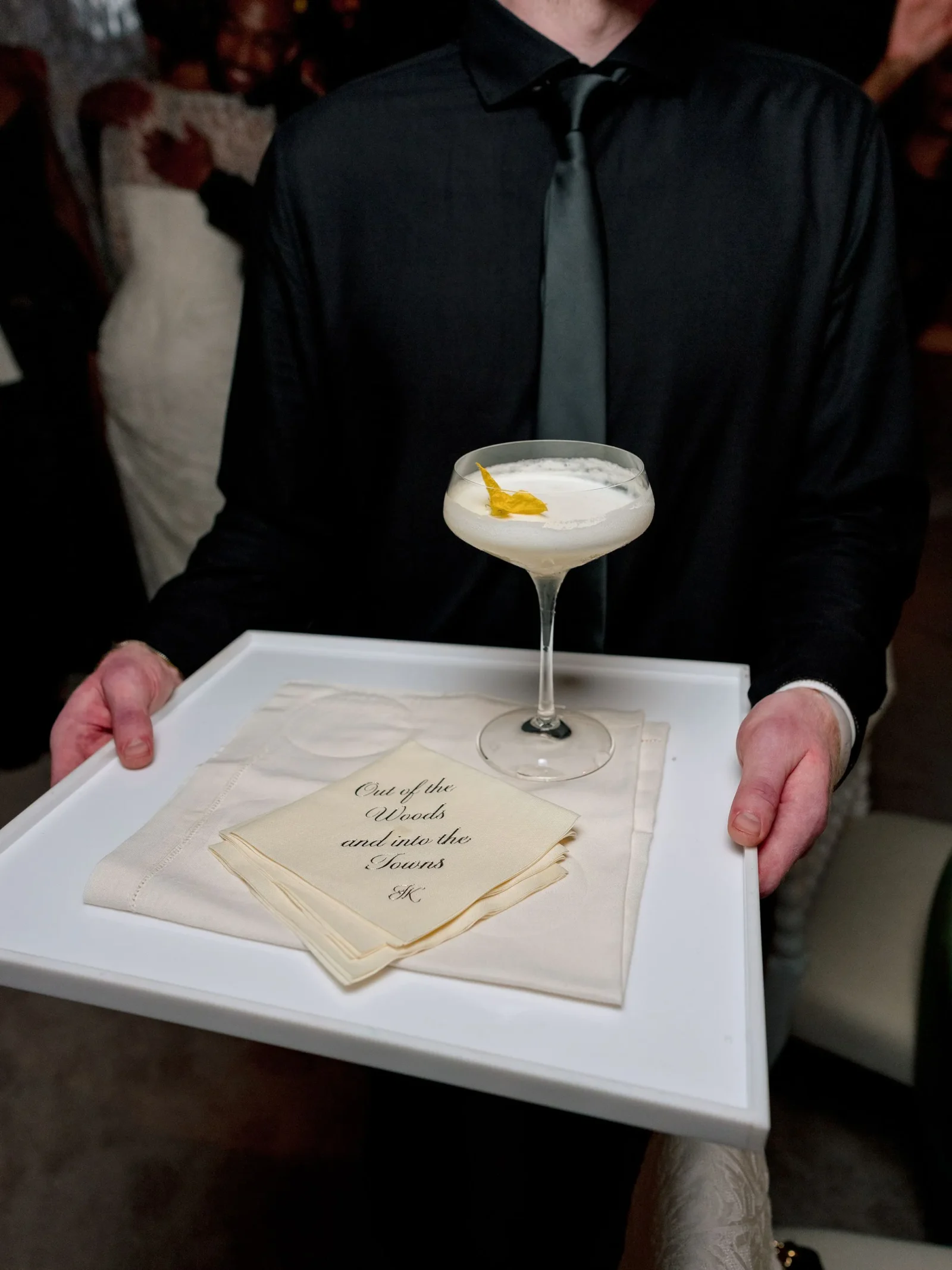 Waiter in a black shirt and gray tie presents a tray with a pale cocktail and napkins.