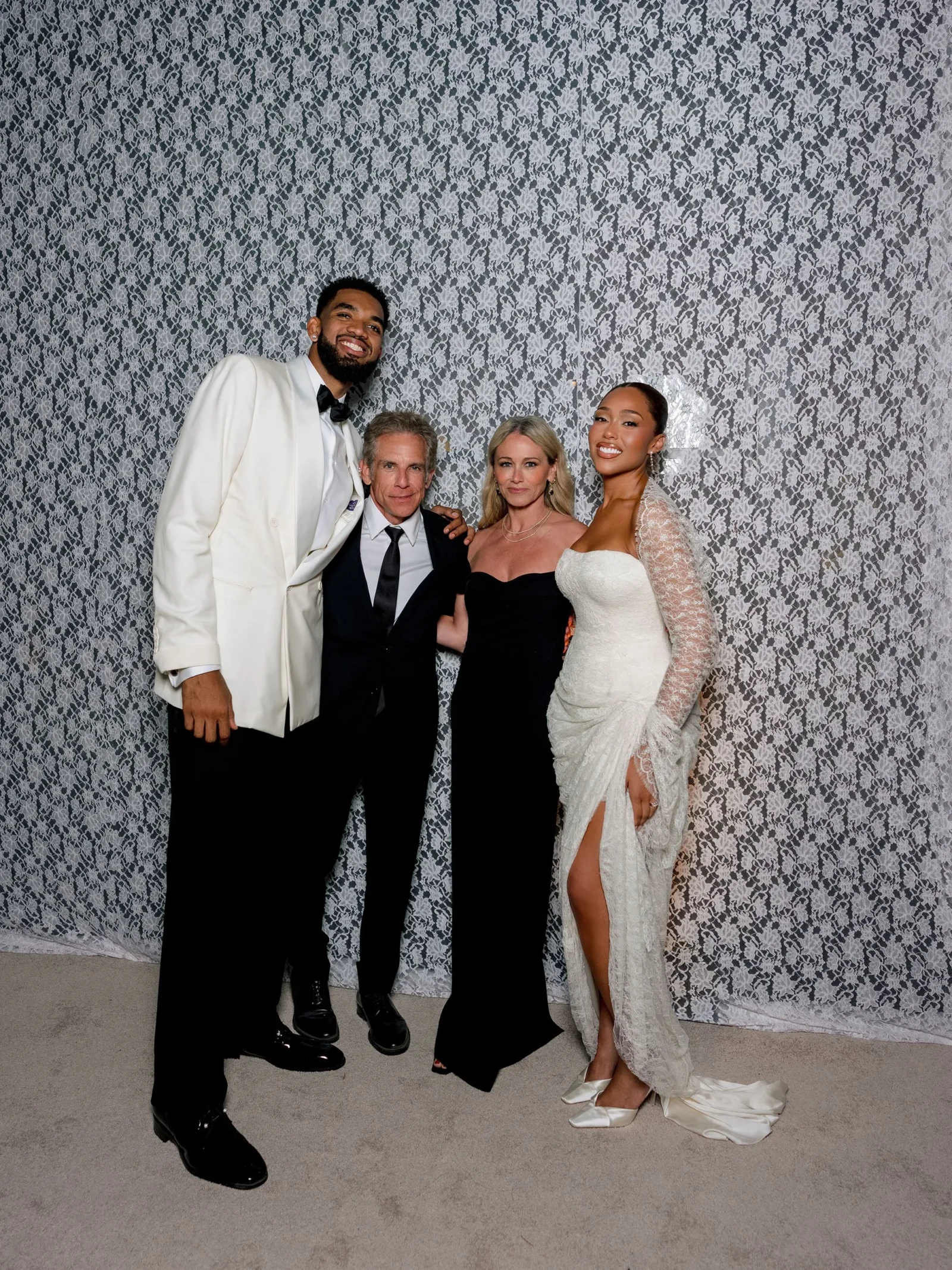 Four people in formal attire pose together in front of a lace-patterned backdrop at a gala or photoshoot.