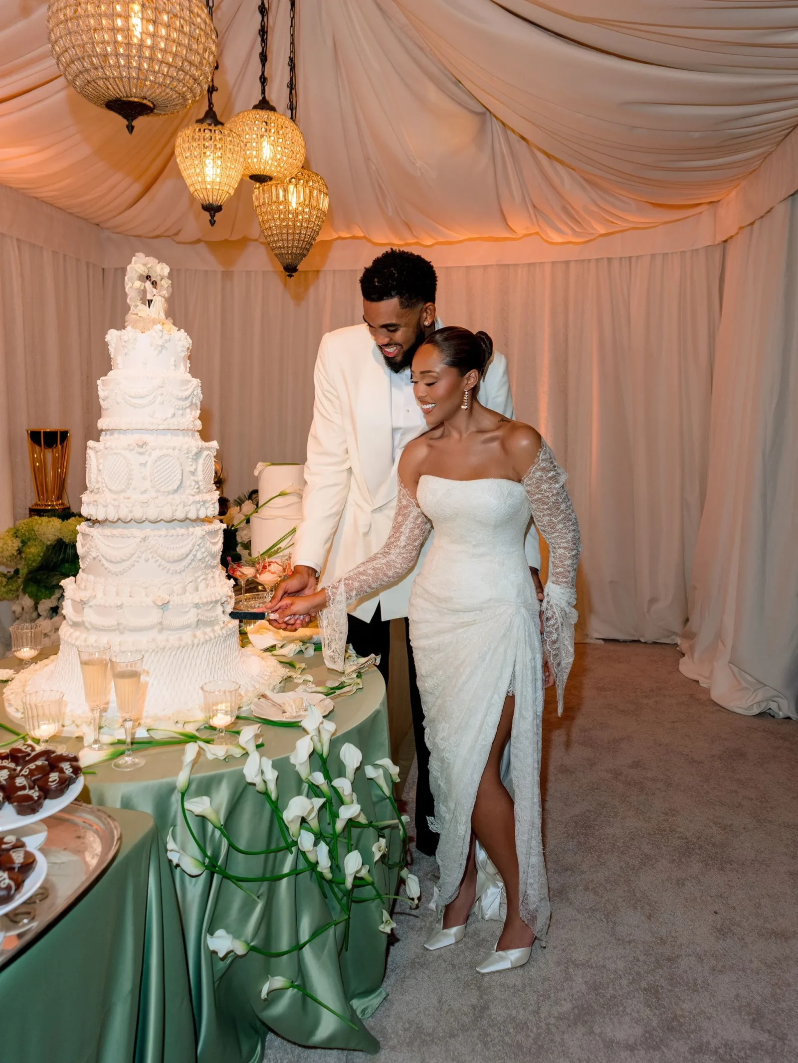 Bride and groom cut a tall white wedding cake under warm chandeliers in a pastel-lit reception hall, smiling at the moment of slicing.