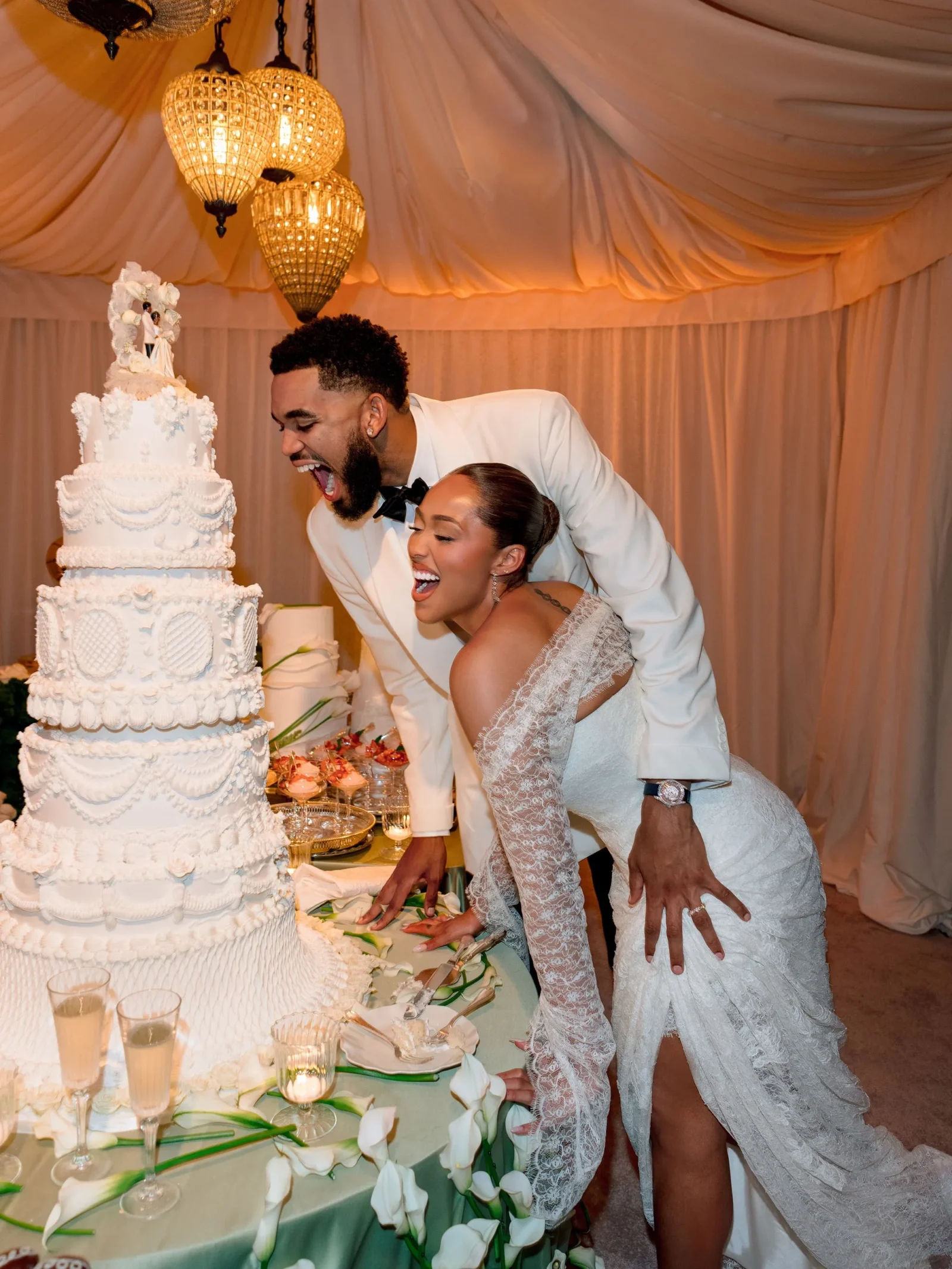 Bride and groom cutting a tall, ornate white wedding cake together at their reception under warm chandeliers.