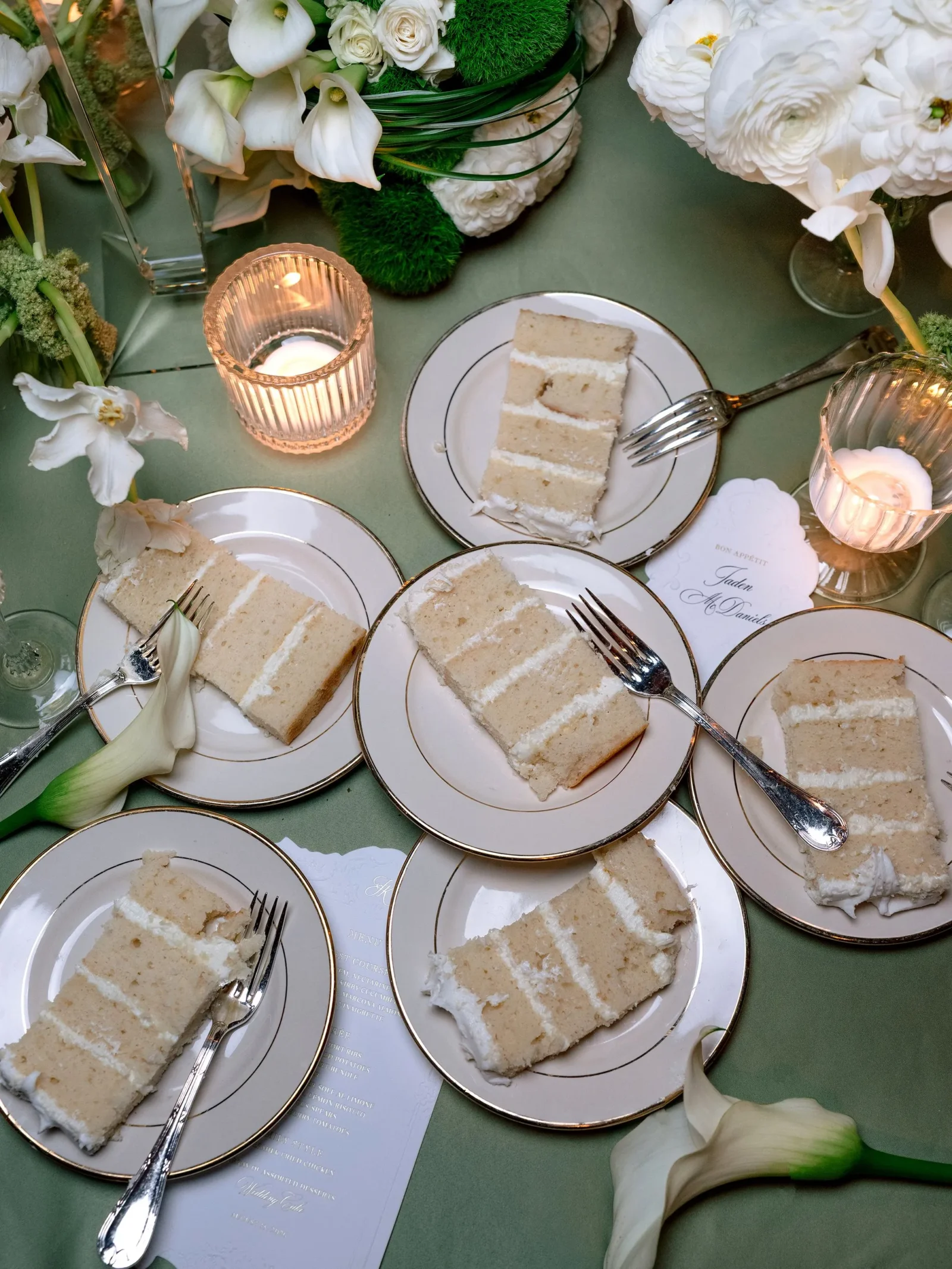 Slices of layered vanilla cake on white china plates with gold rims, surrounded by candles and white flowers on a green tablecloth.