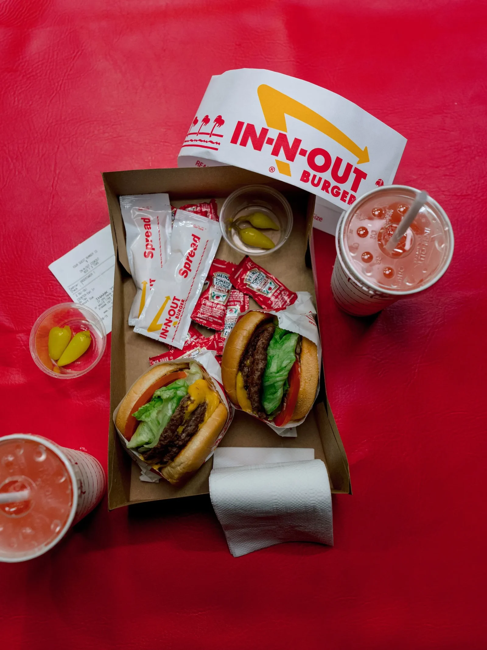 Two In-N-Out burgers in an open box with condiments packets and cups on a red background, showing a casual fast-food meal.