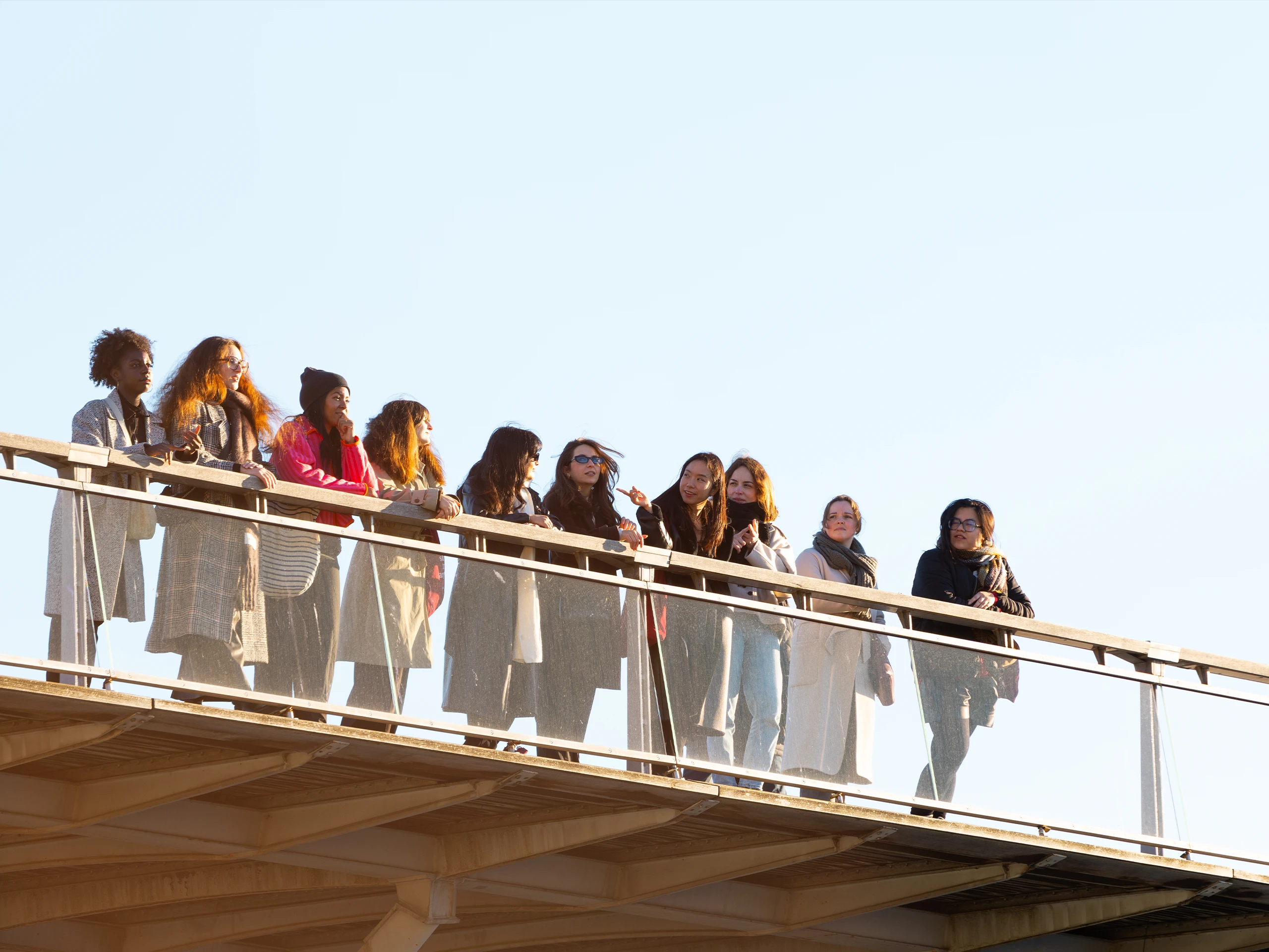 Group of diverse young people leaning on a glass-railed pedestrian bridge, gazing outward at a clear blue sky