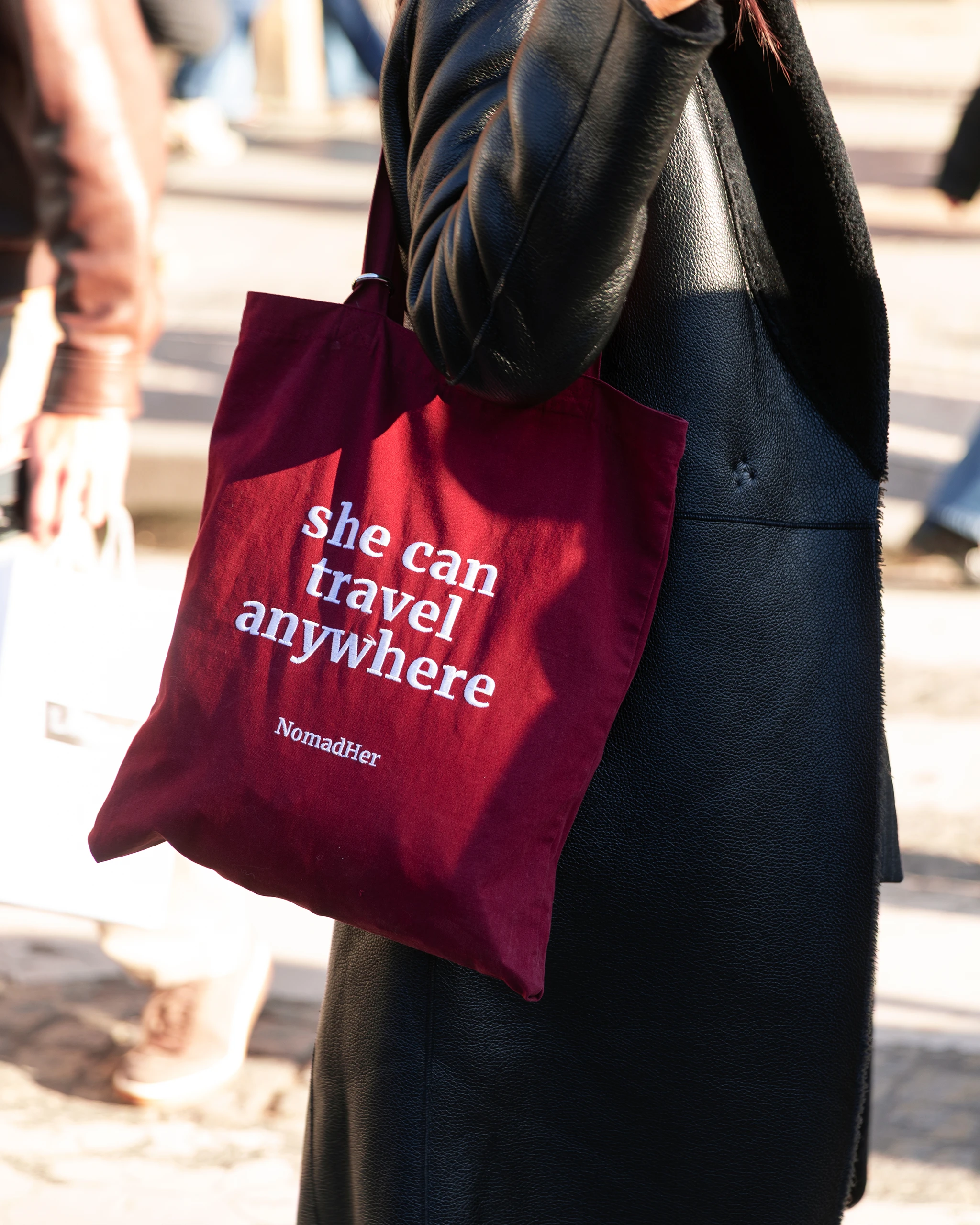 Close-up of a burgundy tote bag with white text reading 'she can travel anywhere' hanging from a black leather coat outdoors.