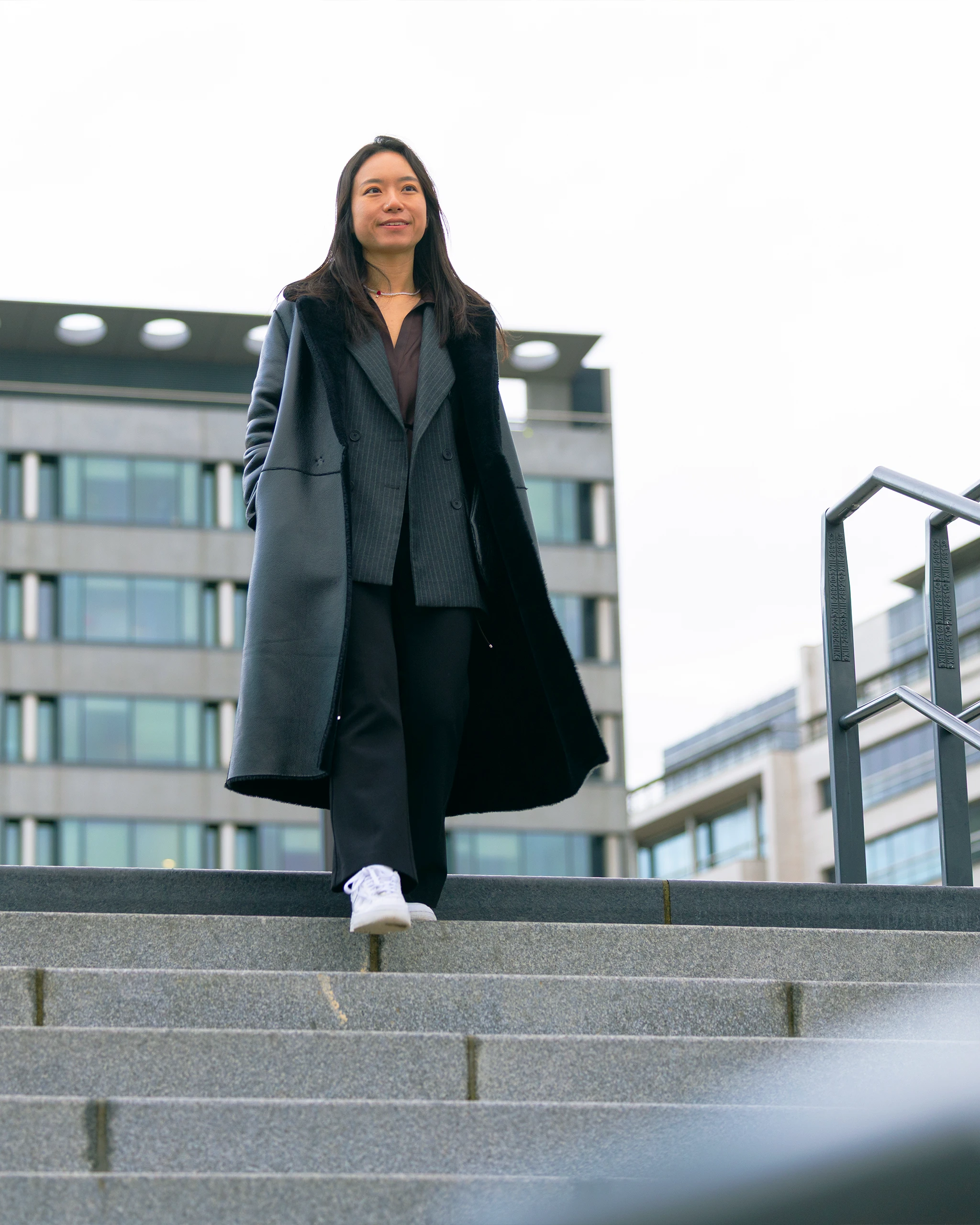 Woman in a dark oversized coat and pinstripe suit stands on outdoor steps in front of a modern glass-front building, looking slightly upward.