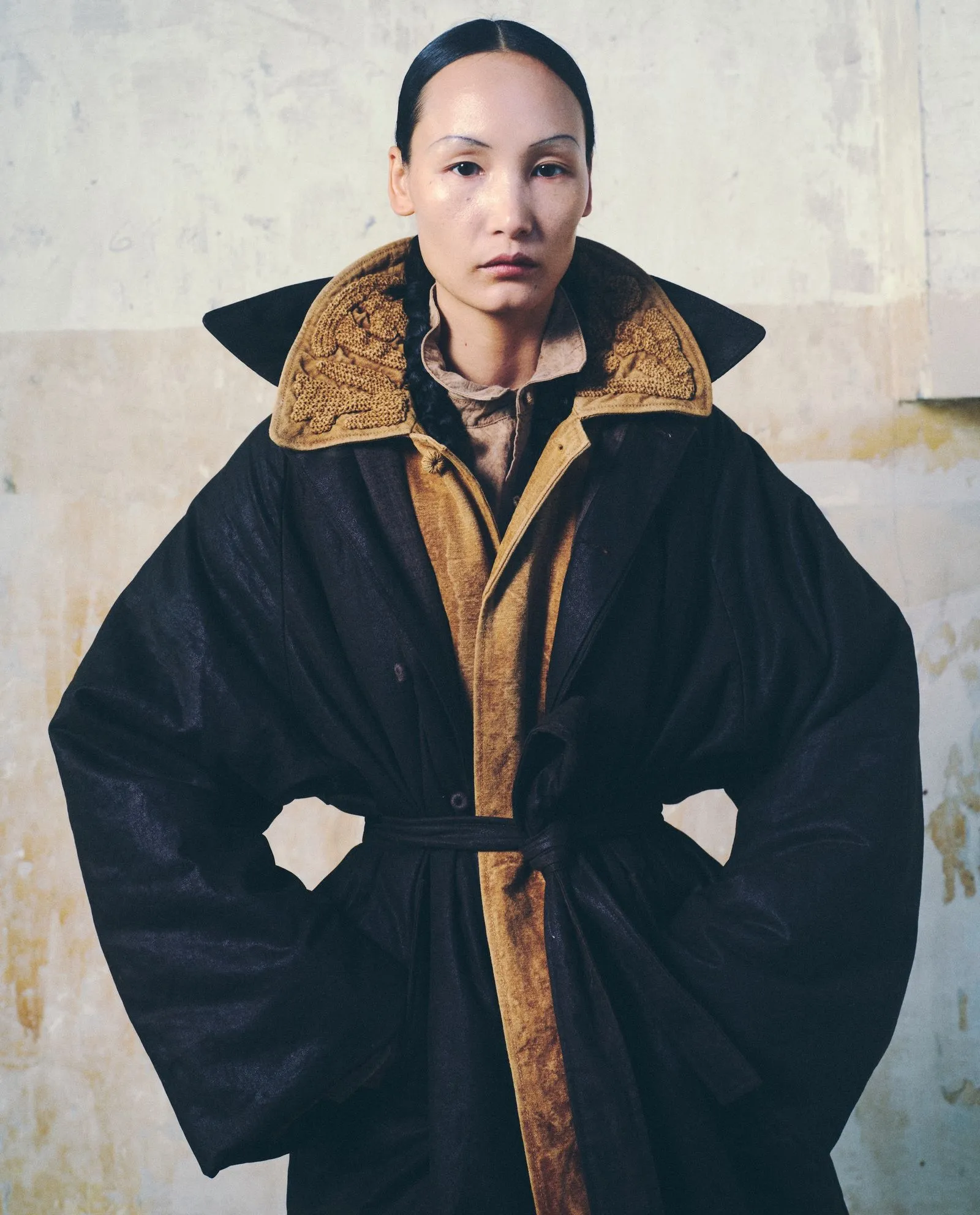 Portrait of a woman facing the camera in an oversized black coat with a brown inner lining and textured lapels, standing with hands on hips against a pale wall.