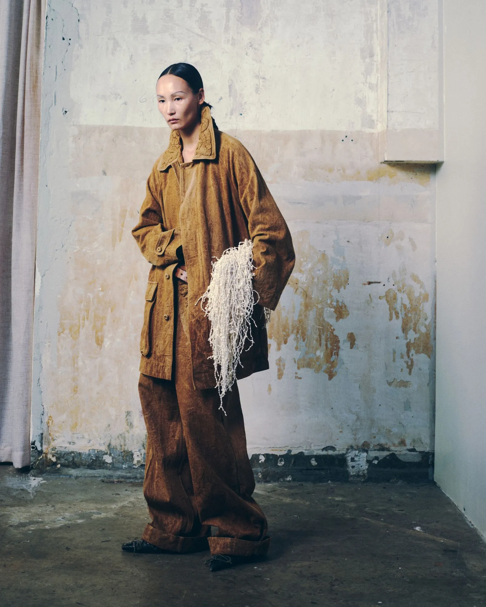 Person in an oversized brown coat stands in a worn, pale-walled room, holding a white tangled netting.