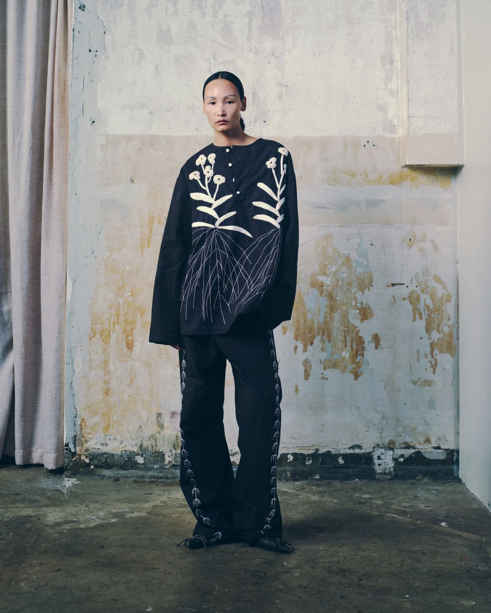 Person wearing a black embroidered outfit with white floral designs, standing against a weathered wall backdrop.