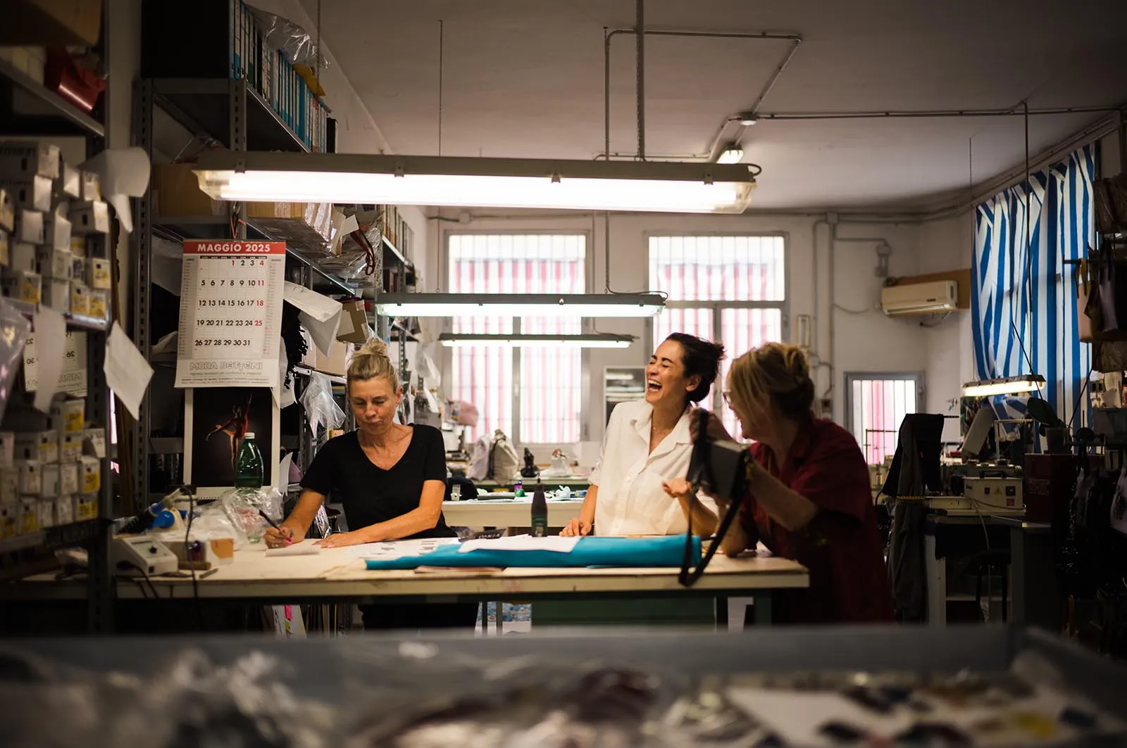 Three women at a large worktable in a busy workshop, drafting and laughing together while shelves clutter the background.