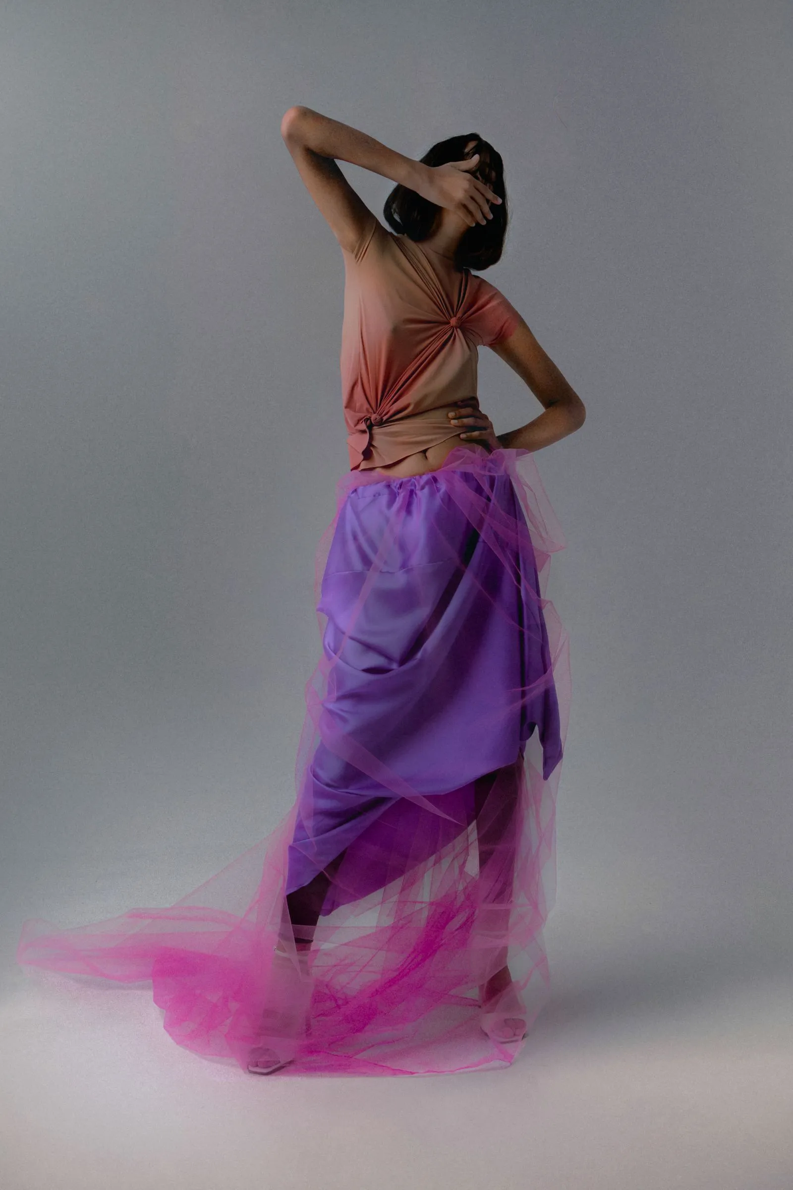Dancer viewed from behind, wearing a peach sleeveless top tied at the back and a purple-pink tulle skirt, posing with one arm bent over the head.