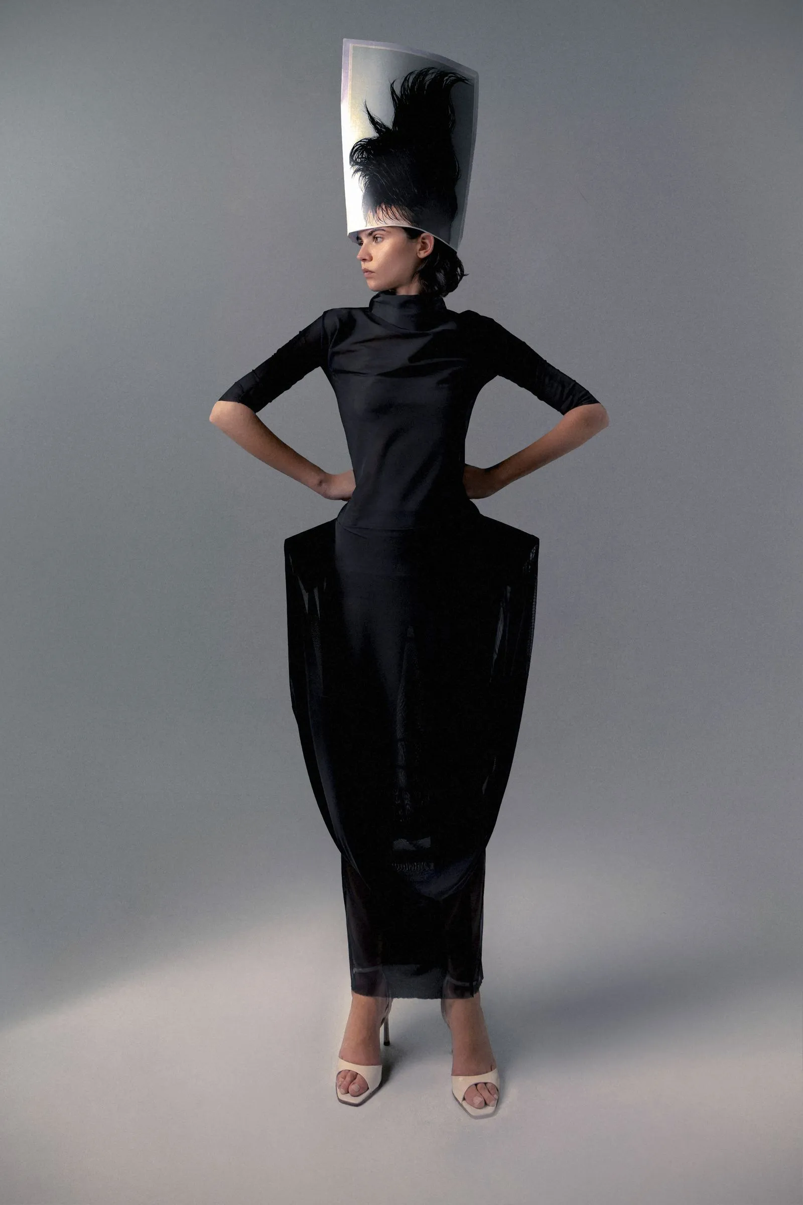 Model in a black, high-neck sheath dress with a large, sculptural gray hat featuring a tall black feather, standing with hands on hips against a gray studio backdrop.