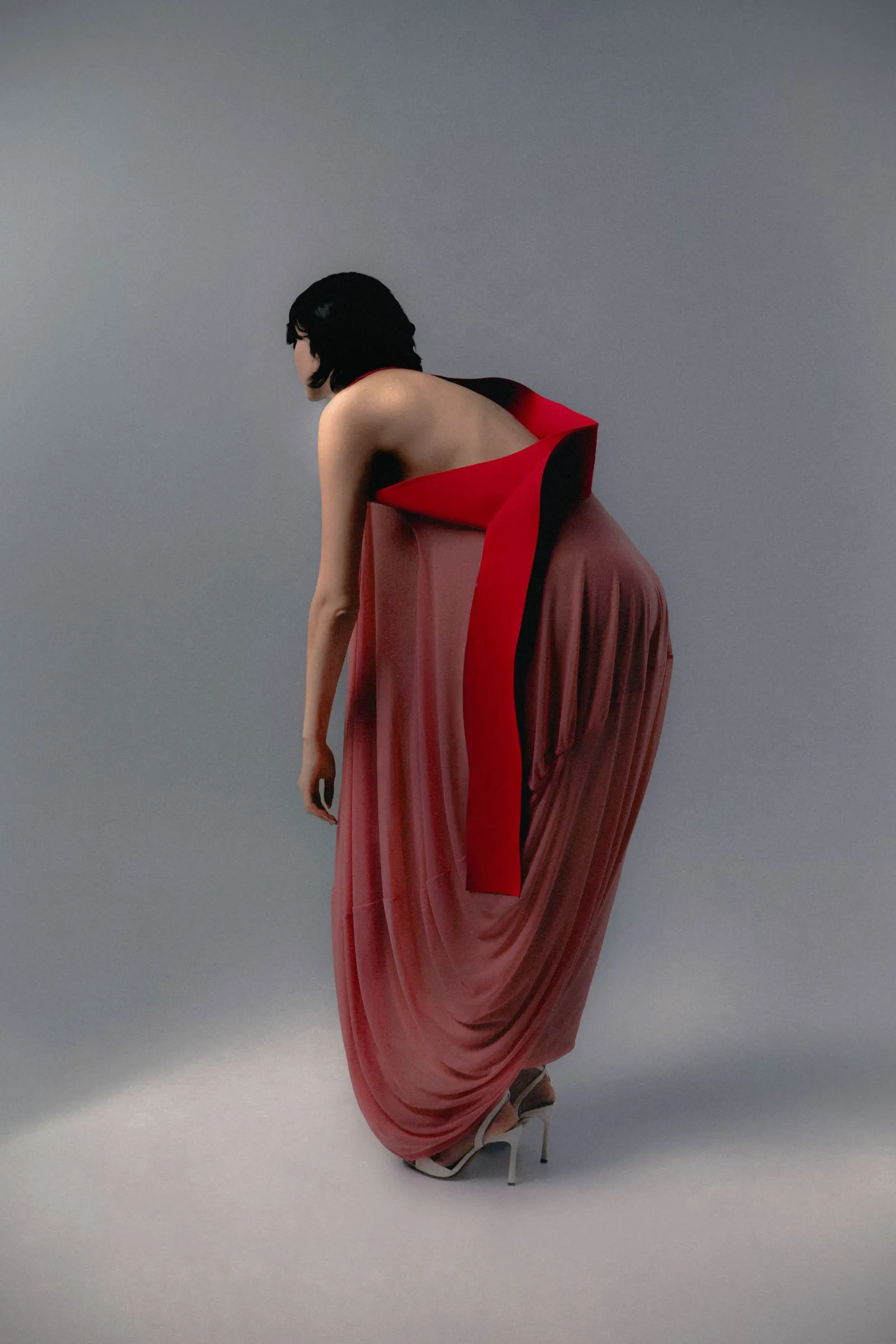 Woman in a pink dress with a bold red sash draped over her back, standing sideways in a studio against a gray backdrop, wearing white heels.