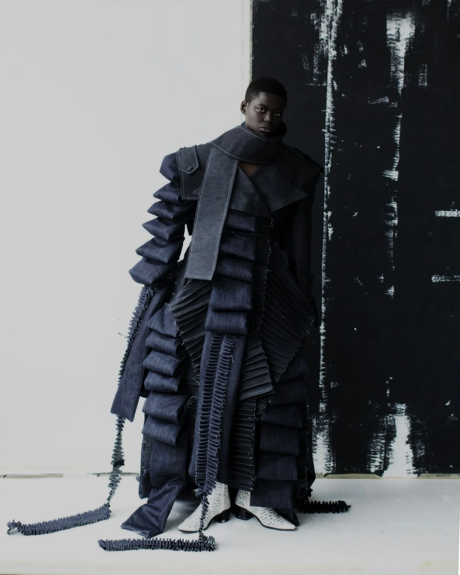 Person in a dramatic, layered dark denim outfit with ruffled panels, standing against a light and dark backdrop.