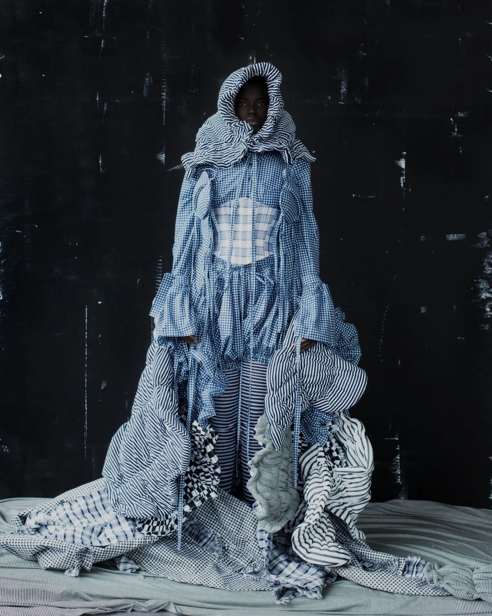Person wrapped in multiple layers of blue-and-white striped fabric standing against a dark wall.