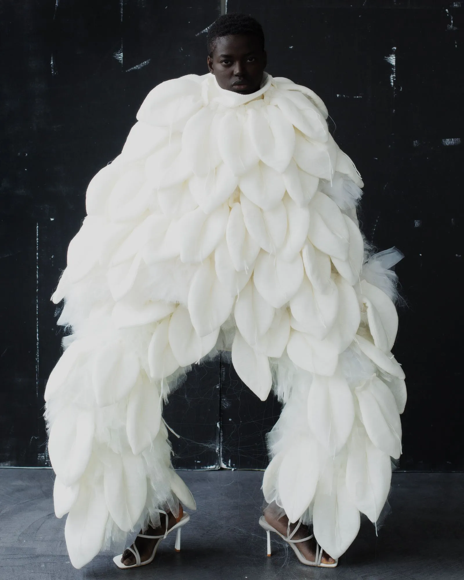 Person in a dramatic white feathered gown with open-toe heels, standing against a dark backdrop.