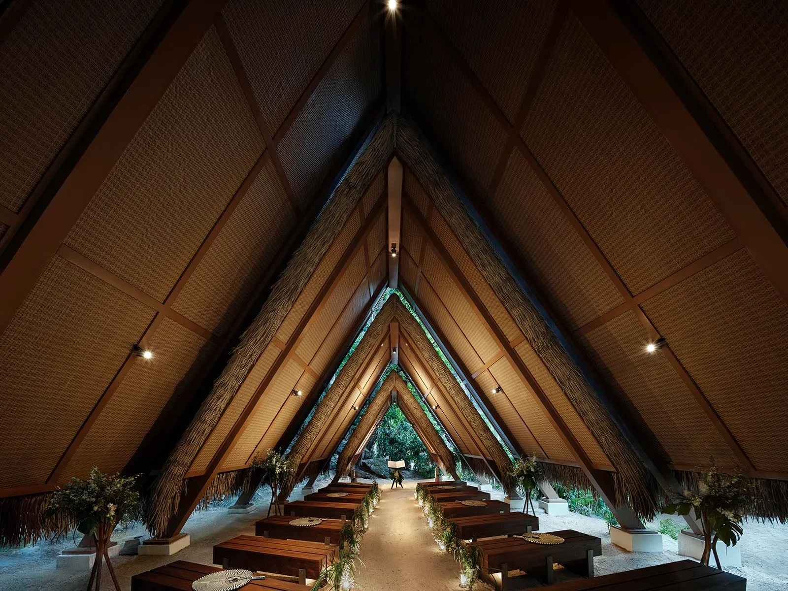 Interior of a thatched, triangular-roof hall with a central aisle, wooden benches, and greenery lining the path for a ceremony.