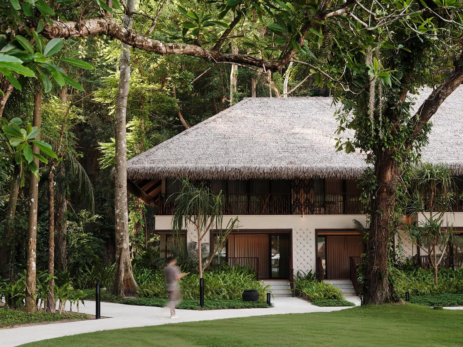 Thatched-roof bungalow nestled in a tropical garden with a curved concrete path and surrounding greenery (blurry passerby in foreground).