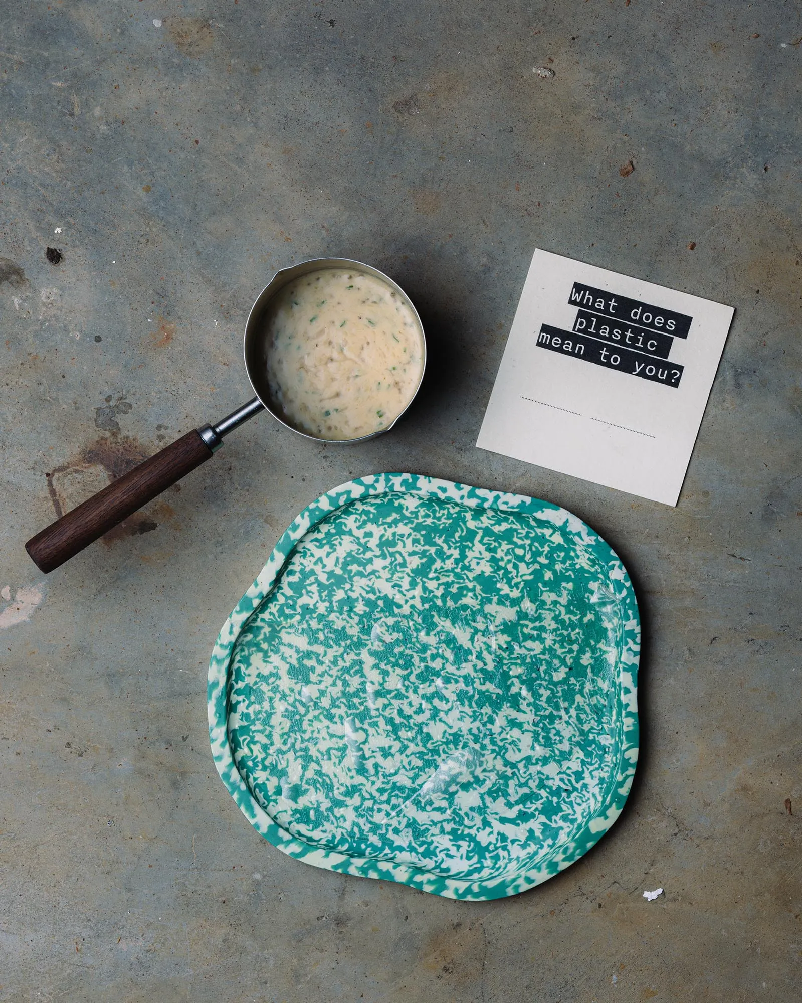 Top-down shot of a saucepan with creamy sauce on a gray concrete floor beside a card that asks, 'what does plastic mean to you?' - with a turquoise patterned plate nearby