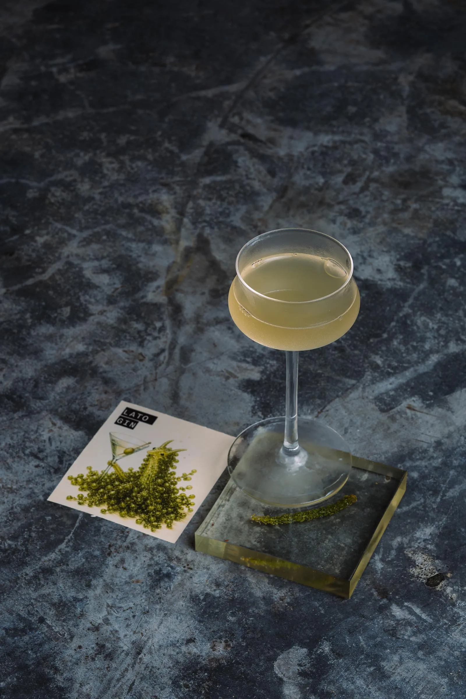A stemmed cocktail glass with a pale yellow drink on a dark marbled surface, beside a botanical illustration card labeled Lato Gin and a small garnish tray.