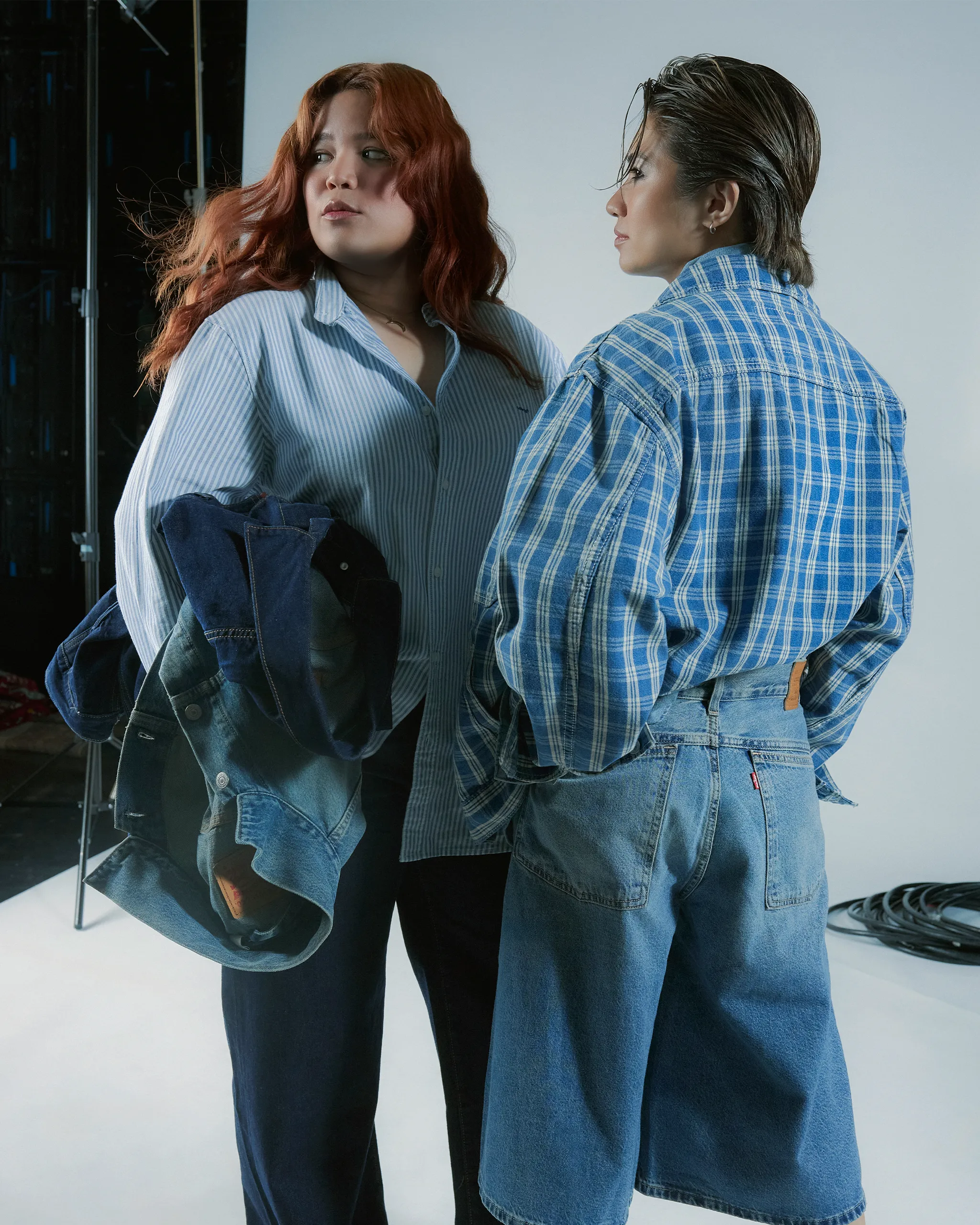 Two people in denim outfits stand back-to-back in a studio, facing opposite directions; one with red hair in a striped shirt, the other with slicked-back hair in a blue plaid shirt and jeans.